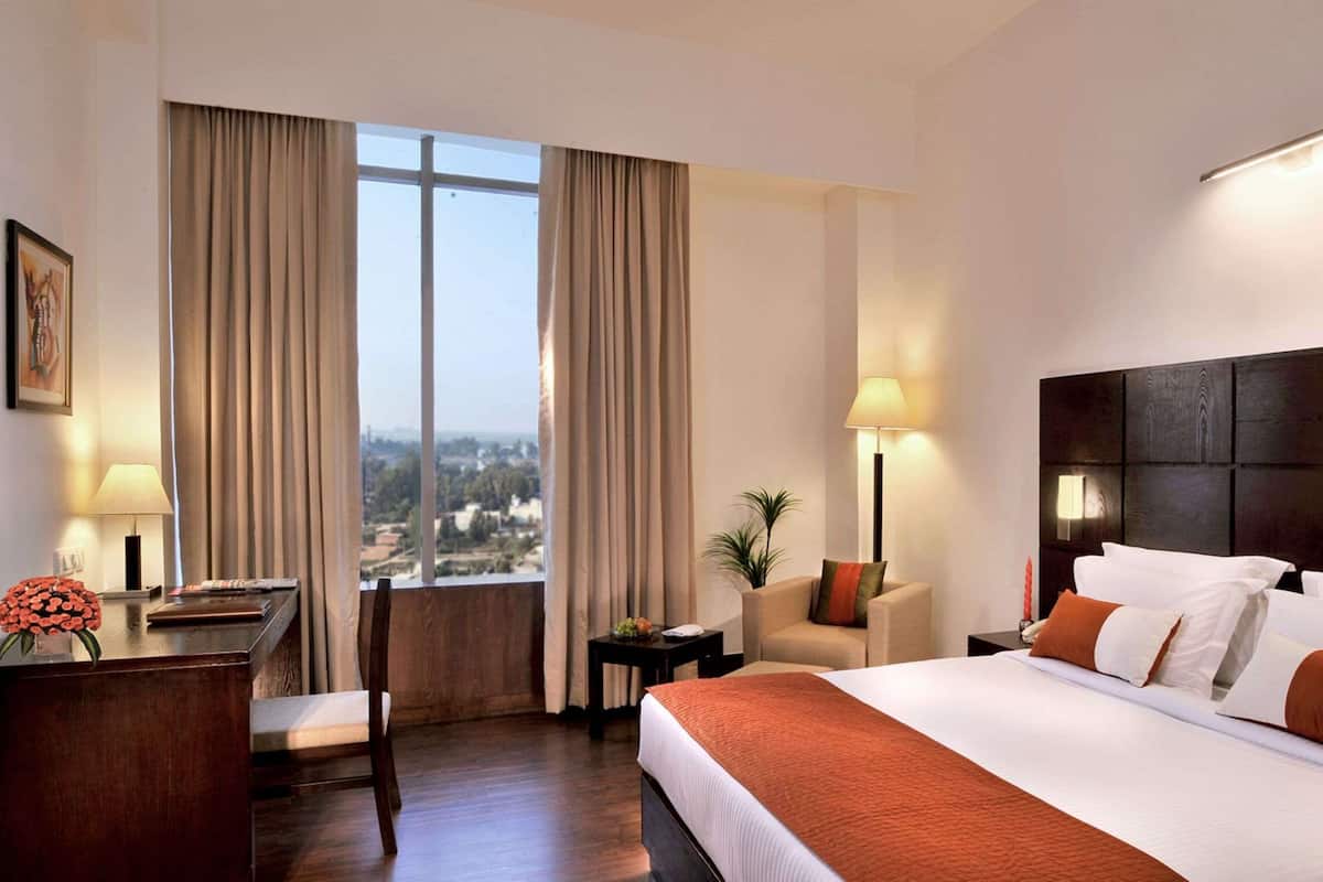 A hotel room with a large bed, a desk, a chair, a nightstand, and a view of the city through large windows.