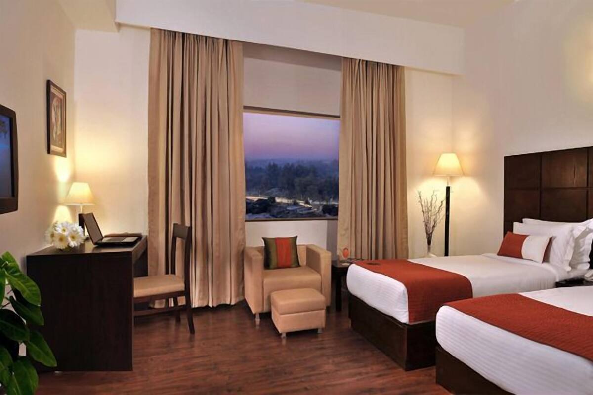 A hotel room with two beds, a desk, a chair, and a large window with a view of a landscape.