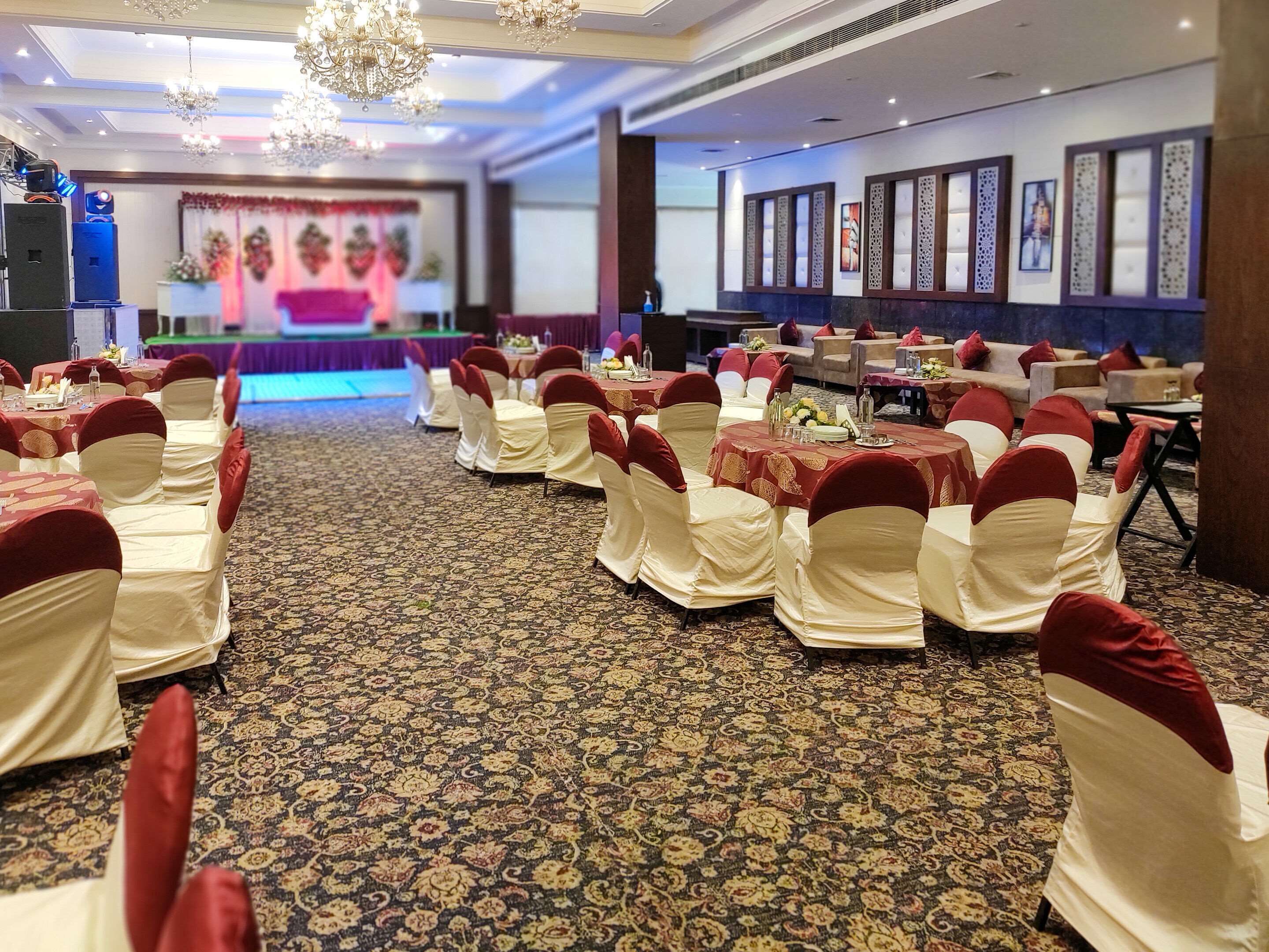 Un salón de banquetes con filas de mesas y sillas, un escenario con una pantalla y paneles decorativos en las paredes.