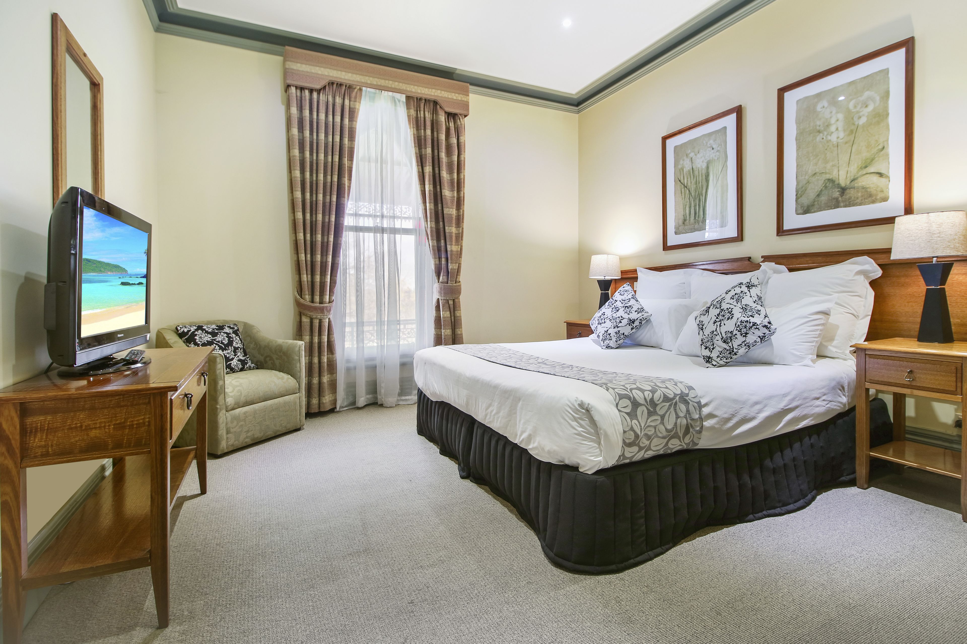 A hotel room with a bed, a desk with a television, a chair, and a window with curtains.