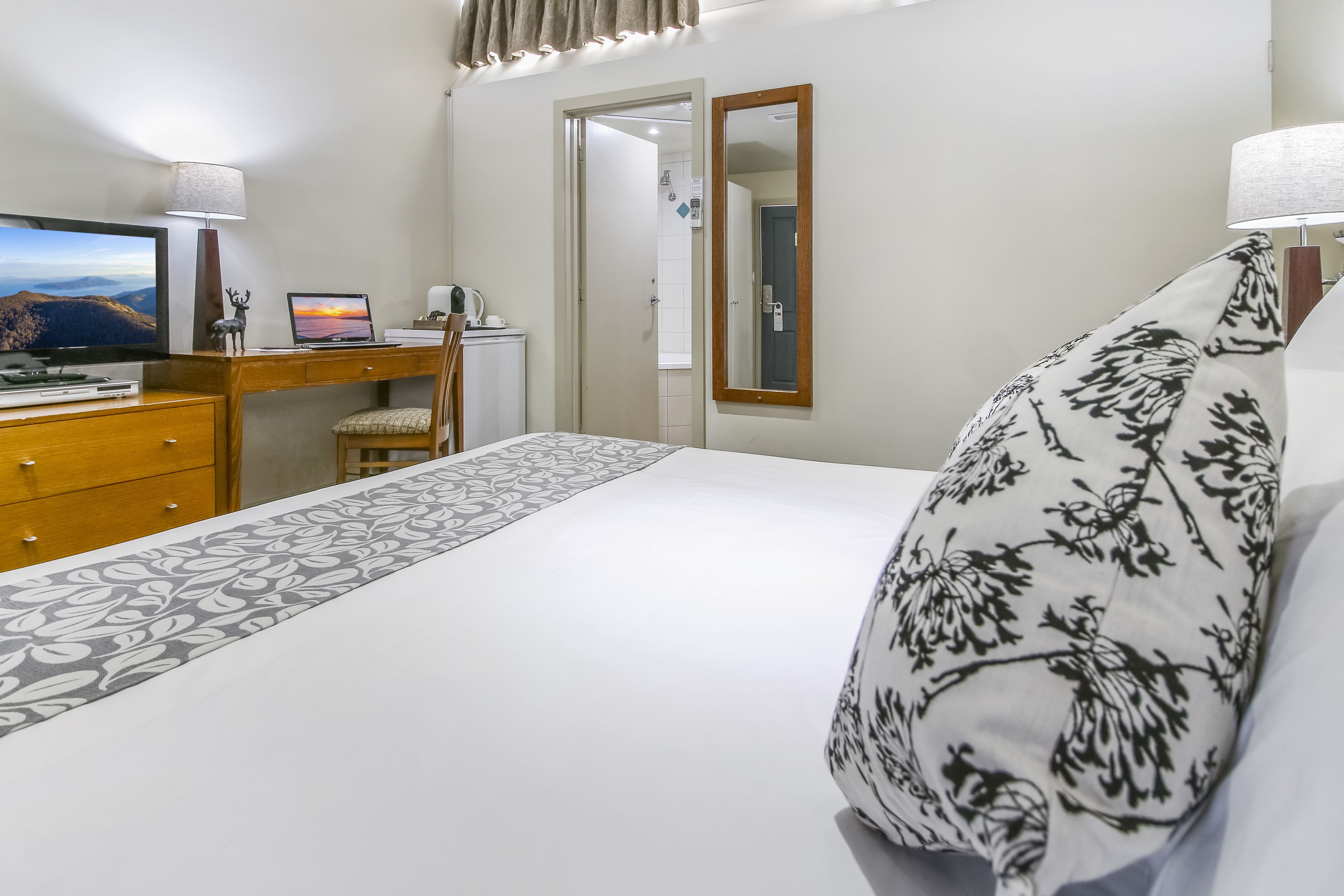 A hotel room with a bed, a desk with a laptop, a TV, and a mirror.