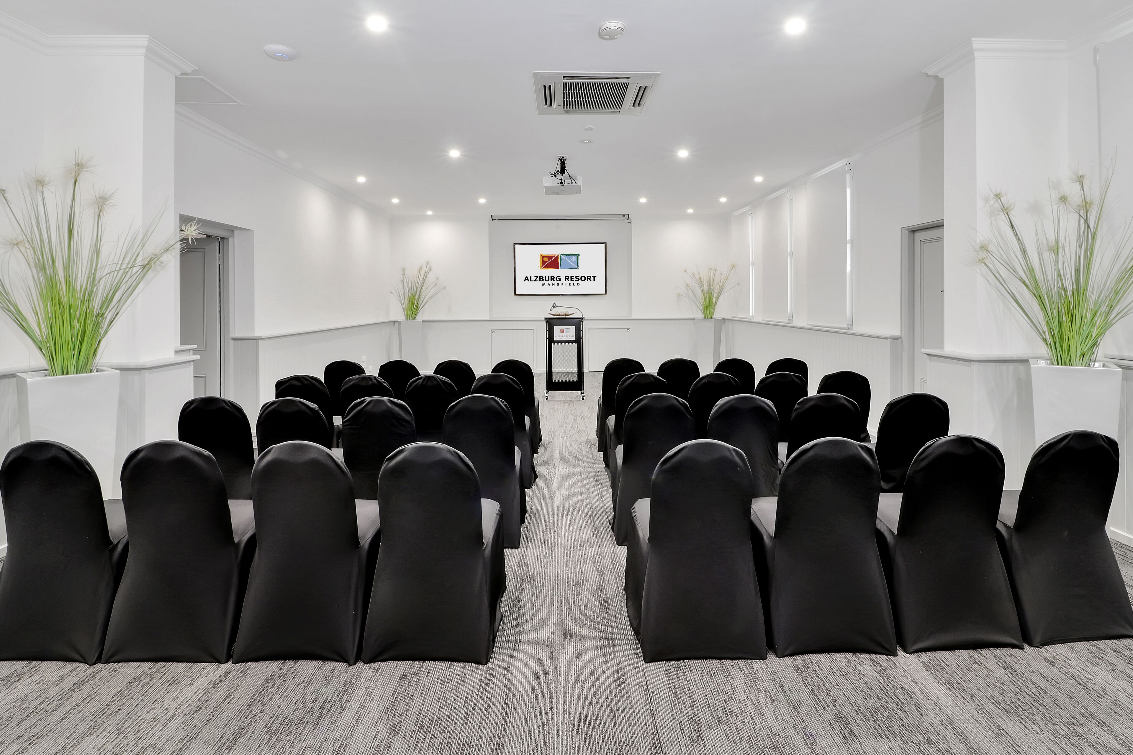 A conference room with rows of black chairs, a projector screen, and a logo for Alzberg Resort.