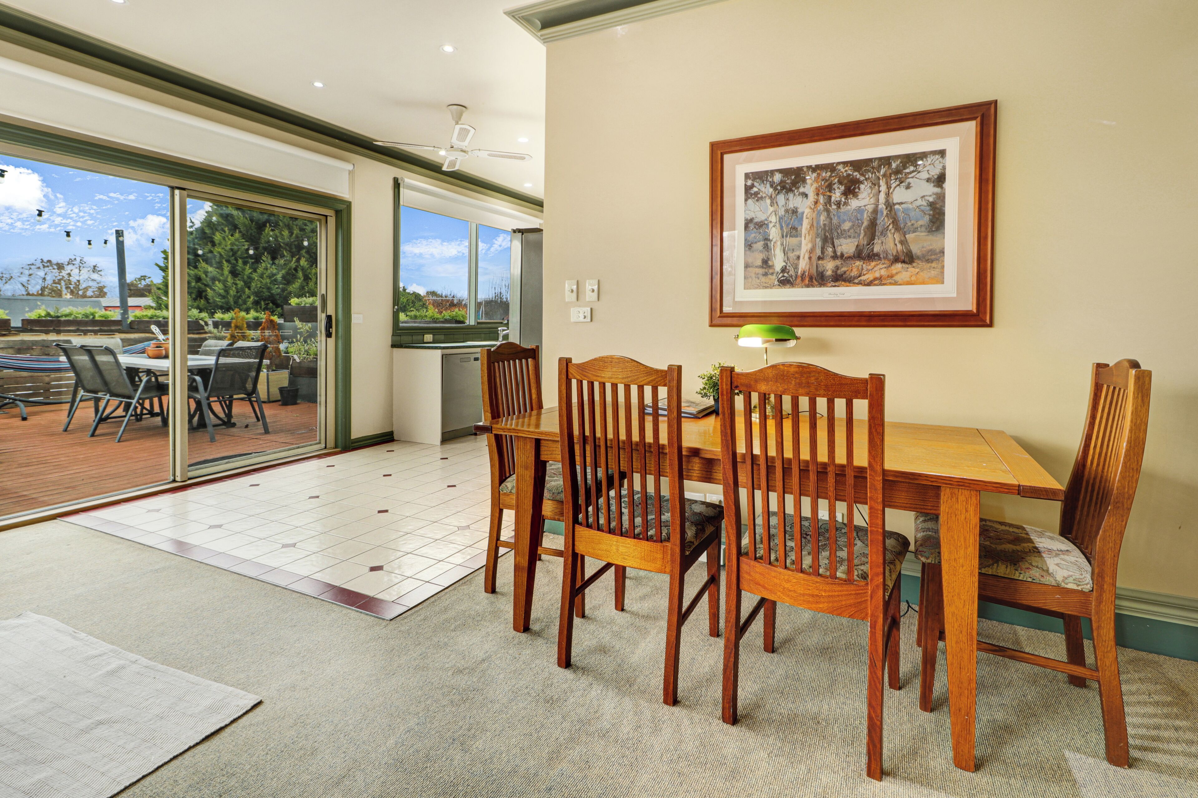 Uma área de refeições com mesa e cadeiras de madeira, um quadro na parede e vista para um pátio externo através de uma porta de vidro deslizante.