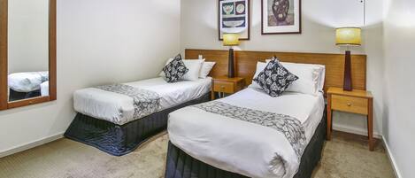 Quarto de hotel com duas camas, cabeceira de madeira, mesa de cabeceira com abajur e um espelho.