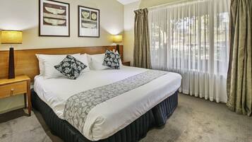 Habitación de hotel con una cama grande, dos lámparas de noche, un cabecero de madera y una ventana con cortinas translúcidas.