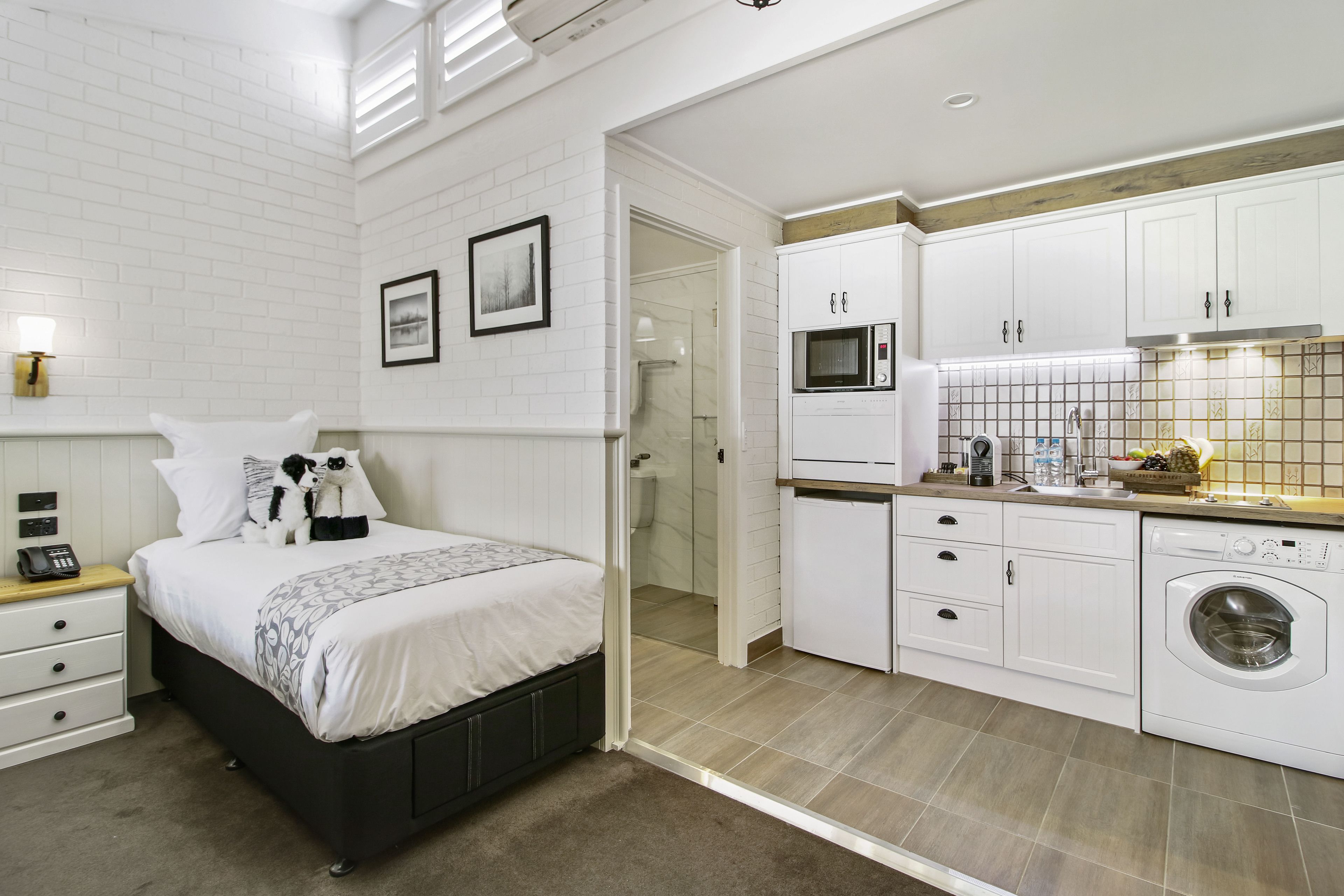 A modern hotel room with a neatly made bed, a small bedside table with a phone, a washing machine, and a kitchenette with a microwave and sink.