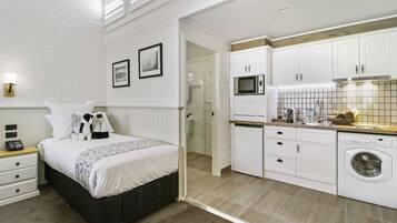 A modern hotel room with a neatly made bed, a small bedside table with a phone, a washing machine, and a kitchenette with a microwave and sink.