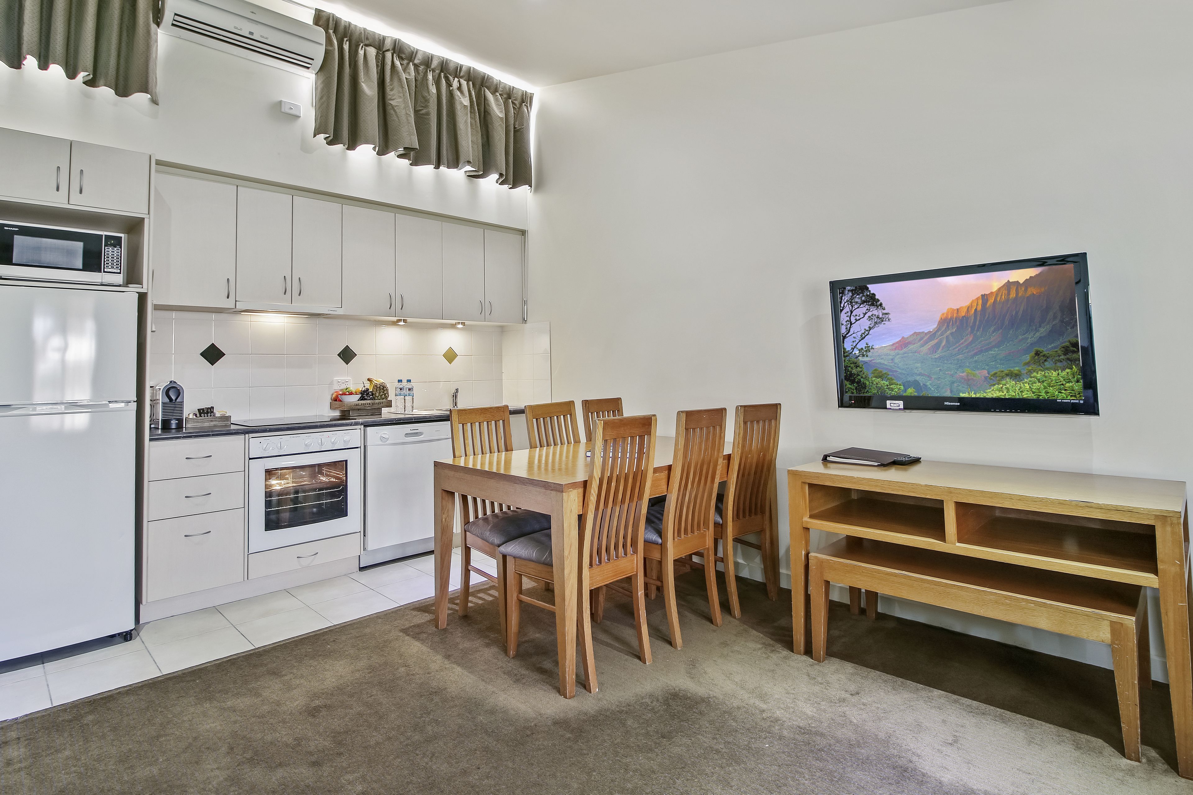 Uma cozinha moderna com armários brancos, micro-ondas e forno. Há uma mesa de jantar com cadeiras e uma TV na parede.