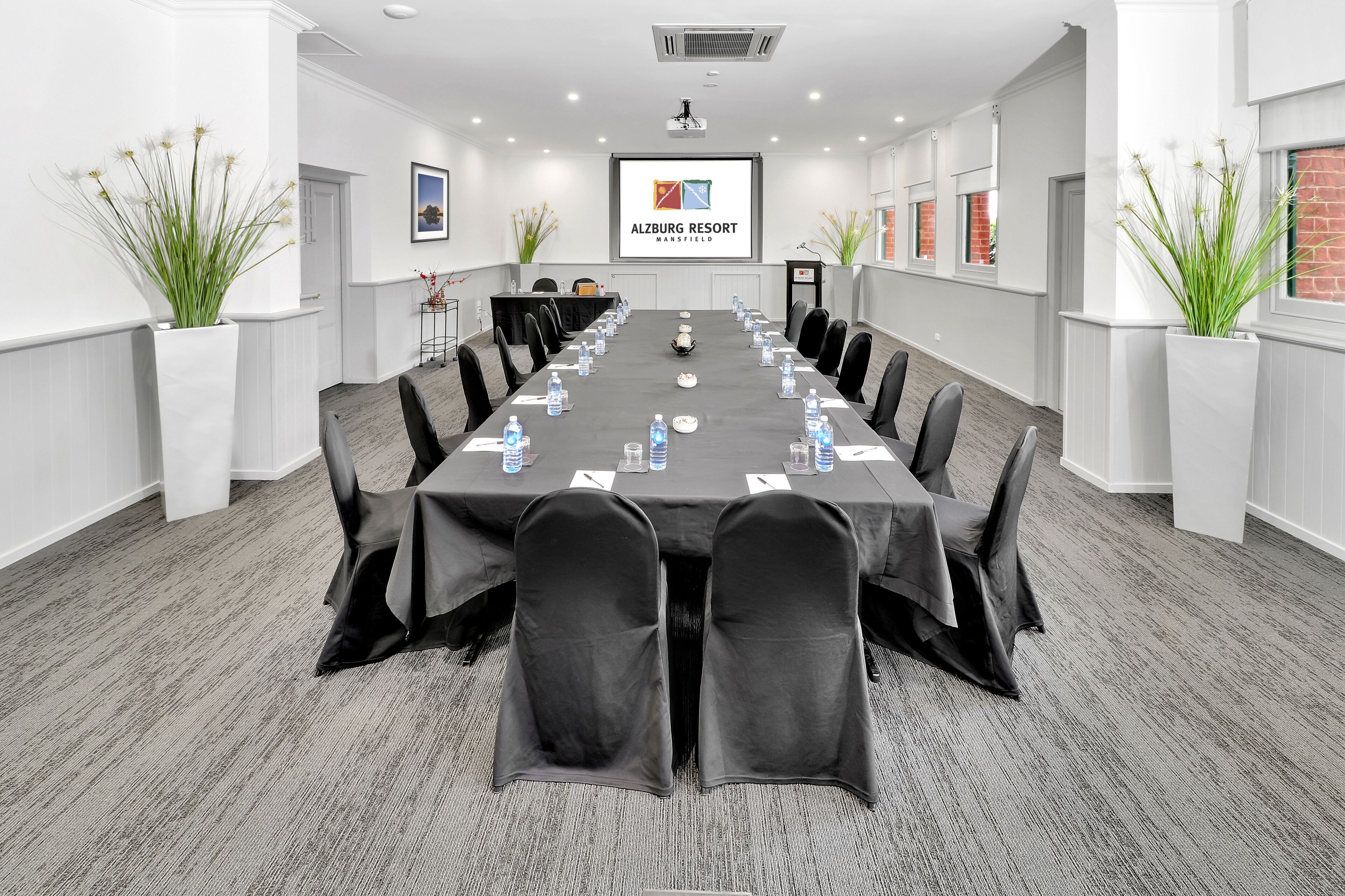 A conference room with a long table, black chairs, water bottles, and a projector screen displaying the logo of Alzberg Resort.