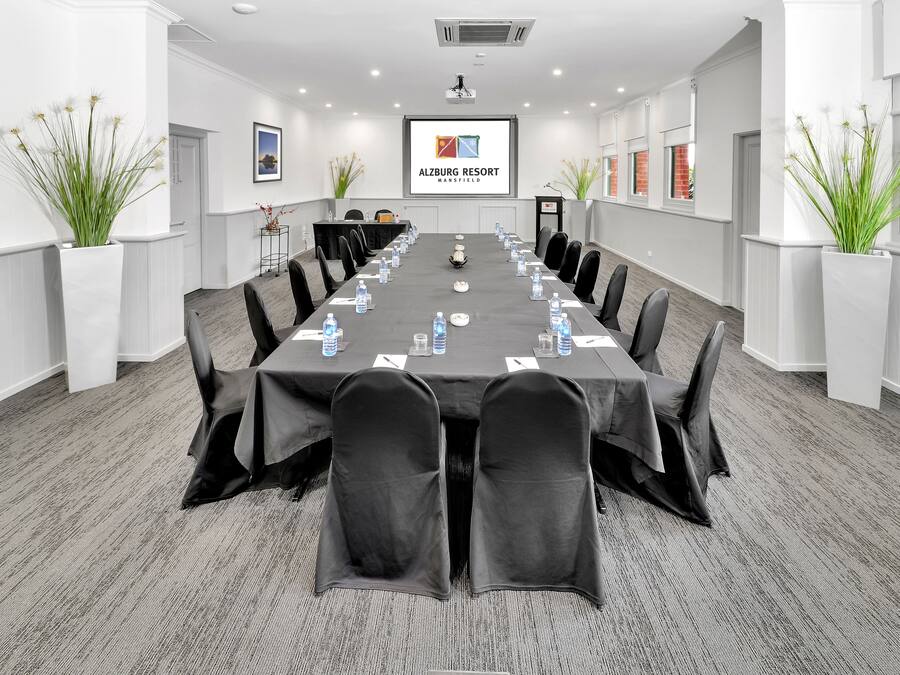 A conference room with a long table, black chairs, water bottles, and a projector screen displaying the logo of Alzberg Resort.