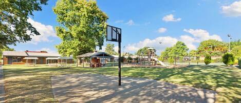 Una cancha de baloncesto con un aro, mesas de picnic y un parque infantil al fondo.