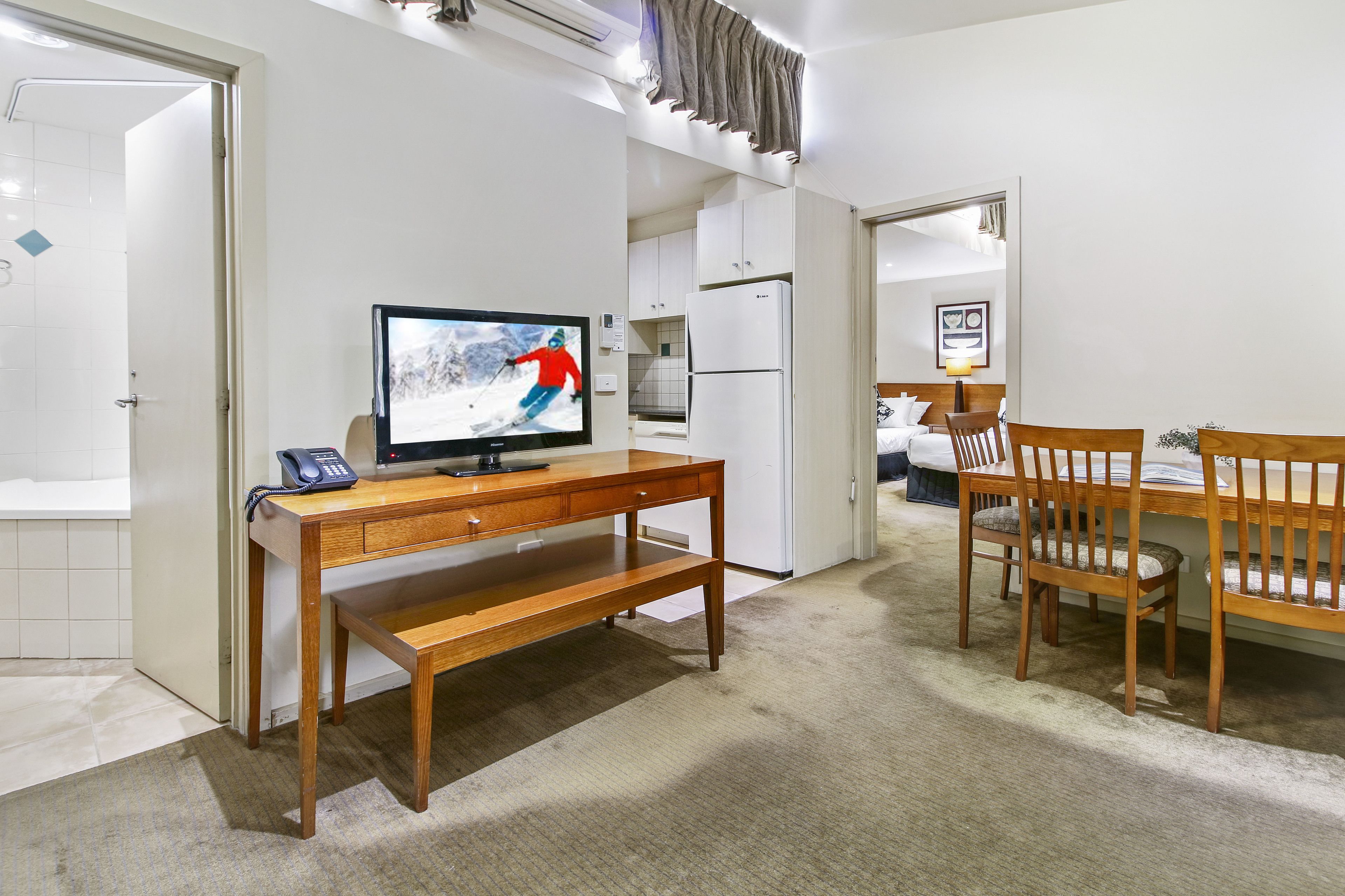 A hotel room with a wooden desk, a television, a refrigerator, a dining table with chairs, and a bathroom visible through an open door.