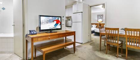 A hotel room with a wooden desk, a television, a refrigerator, a dining table with chairs, and a bathroom visible through an open door.