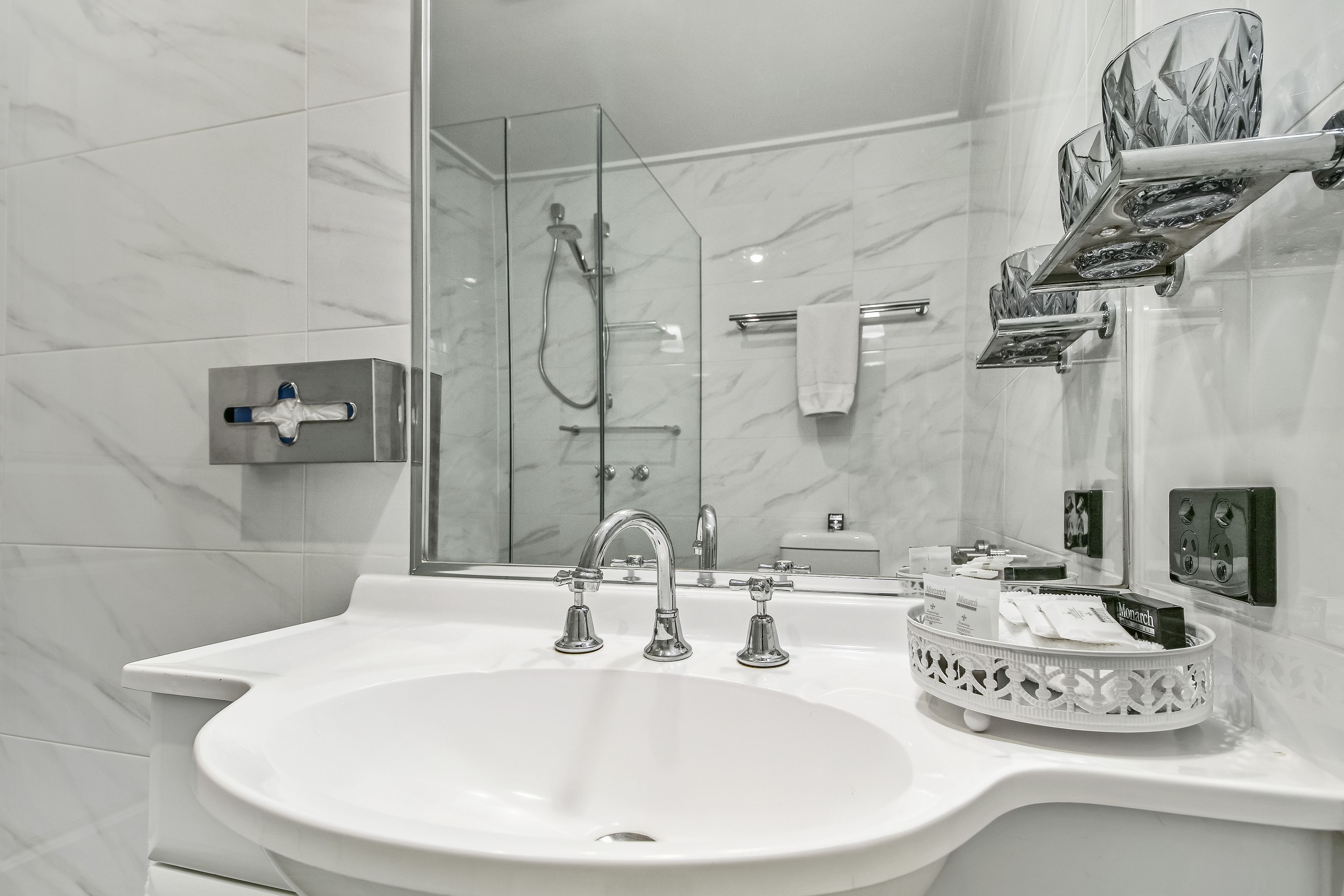 A modern bathroom with a large white sink, a glass-enclosed shower, and a marble-tiled wall.