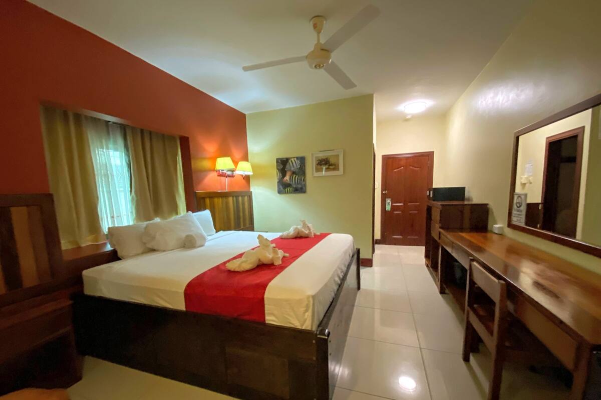 Habitación de hotel con cama, escritorio, silla, ventilador de techo, puerta y ventana con cortinas.