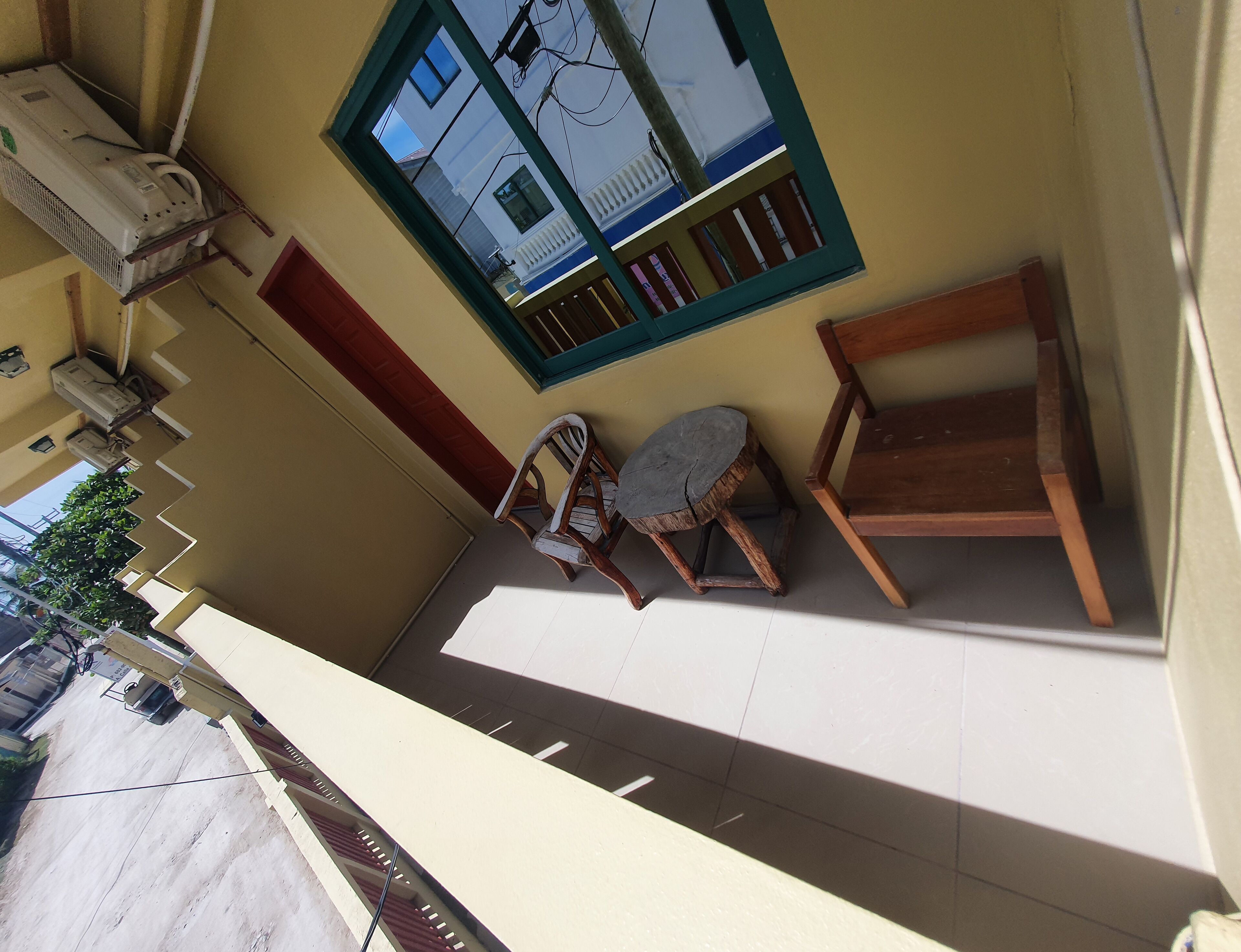 A balcony with a view of a building, a wooden chair, a small stool, and an air conditioning unit.