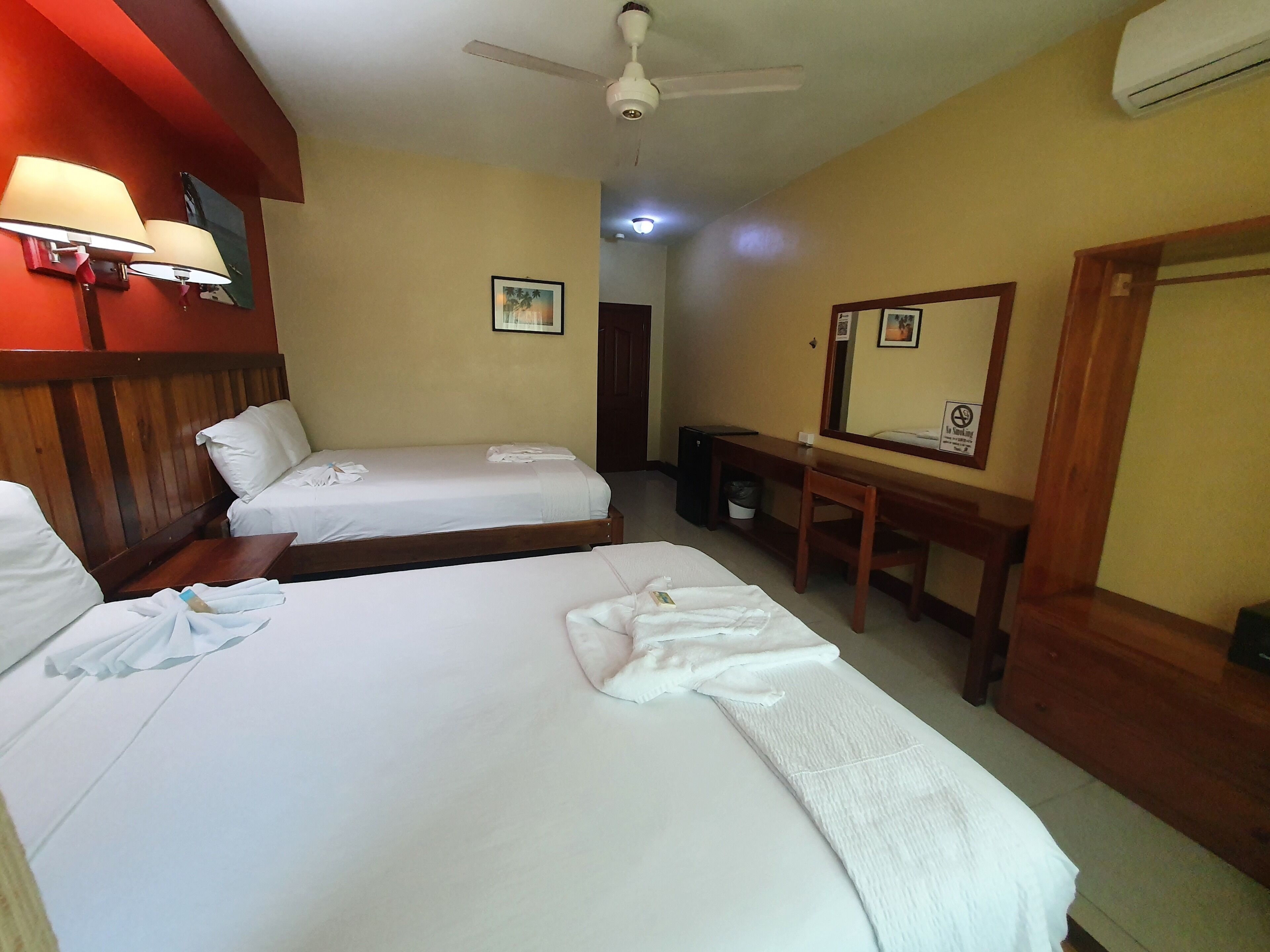 A hotel room with two beds, a desk, a chair, a ceiling fan, and a mirror.