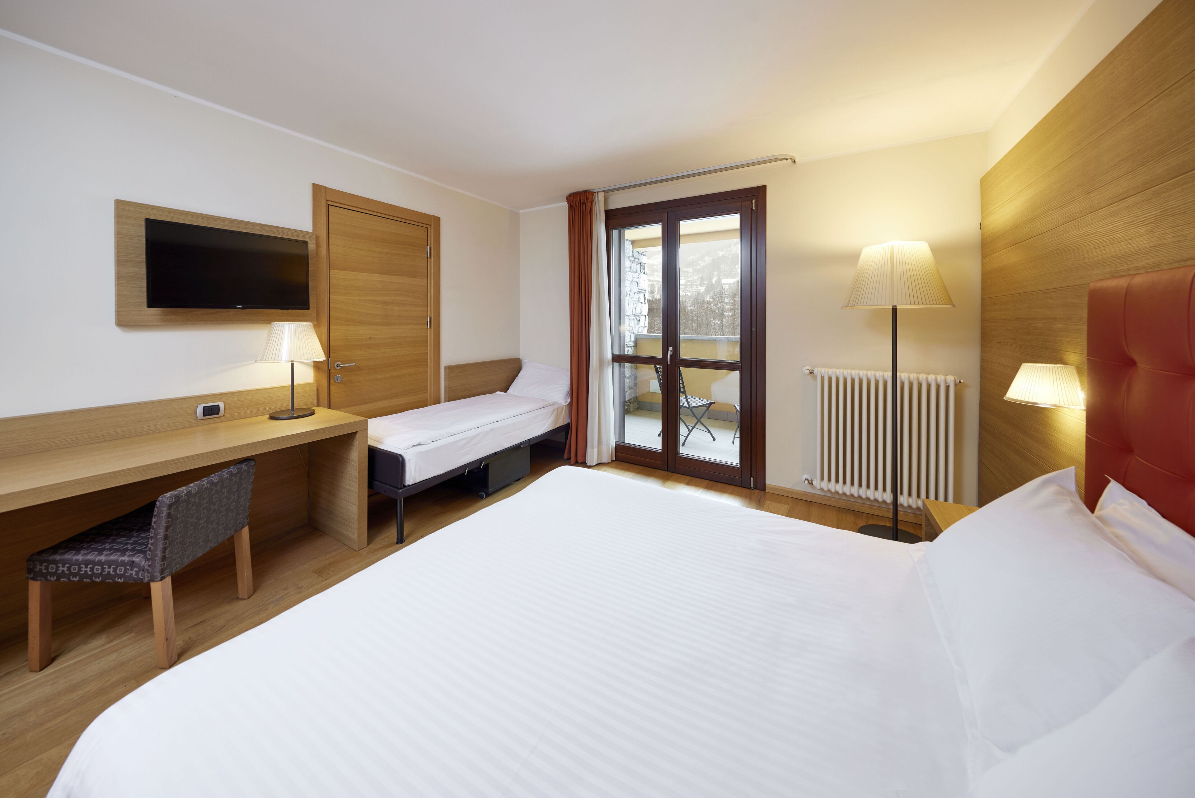 Una camera d'albergo con un letto, una scrivania con una sedia, una TV a parete e una finestra con tende.