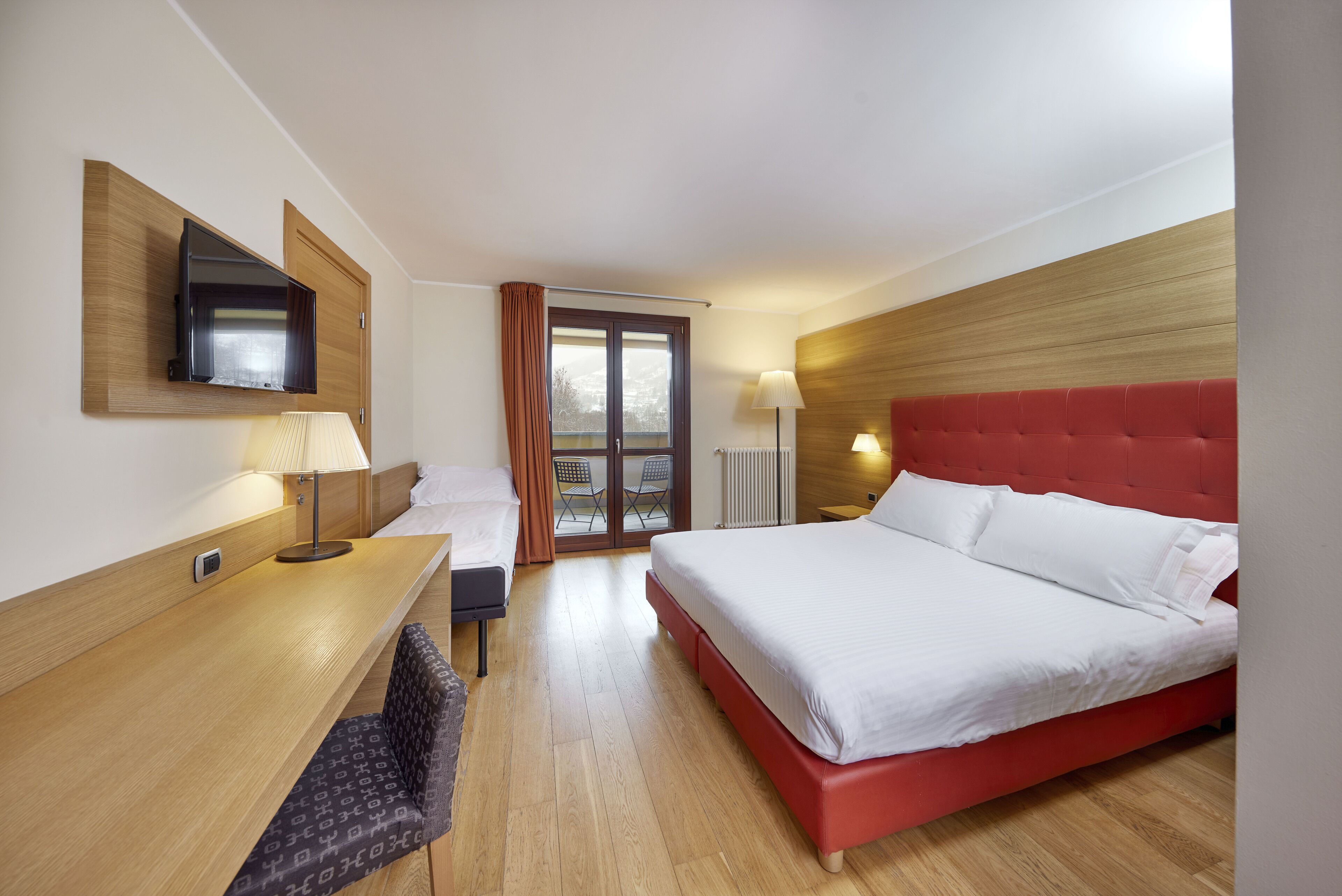 A hotel room with a bed, a desk, a TV mounted on the wall, a window with curtains, and a radiator.