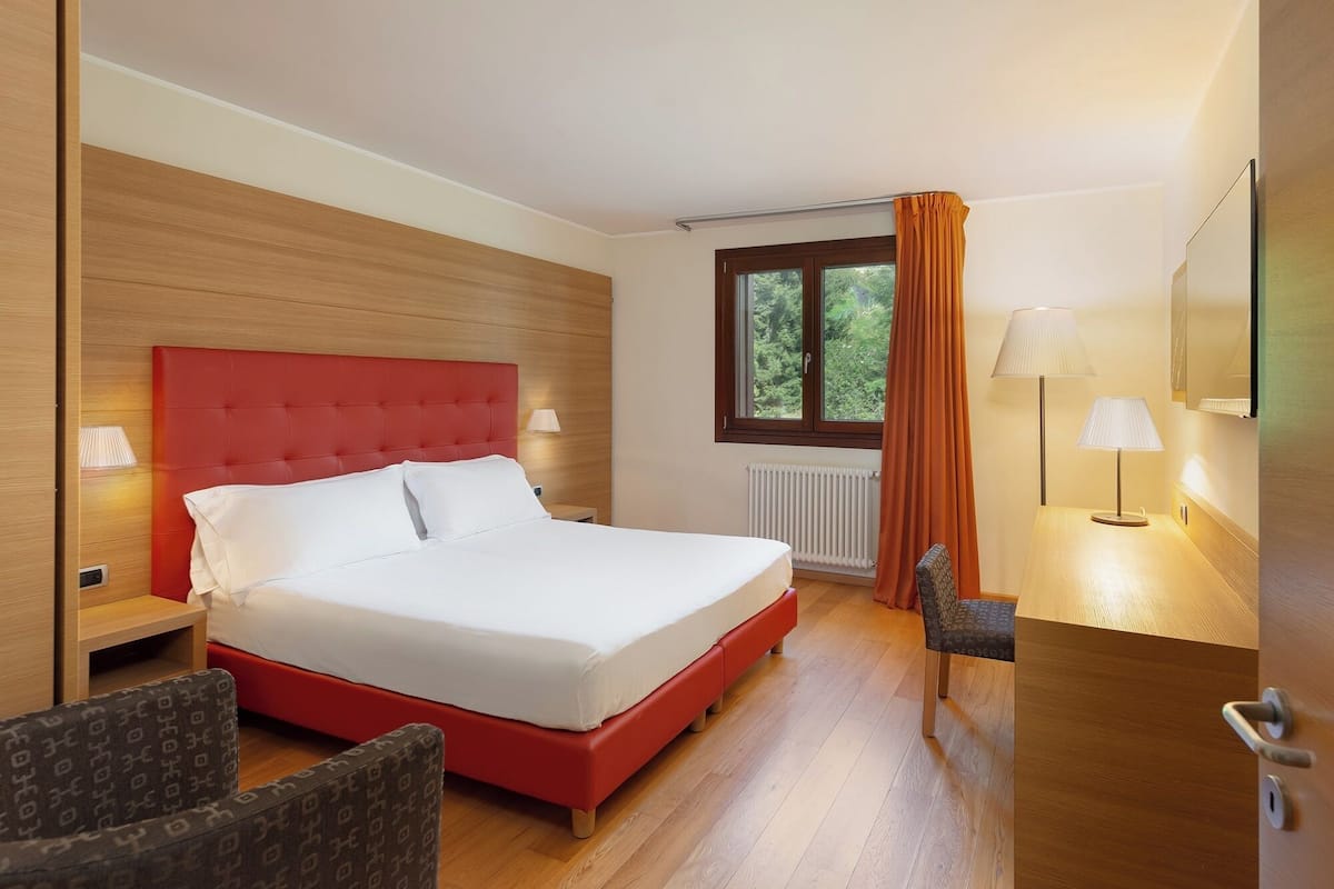 Une chambre d’hôtel avec une tête de lit rouge, un grand lit, deux lampes de chevet, une fenêtre avec des rideaux, un radiateur, un plancher en bois et une petite table avec une lampe.