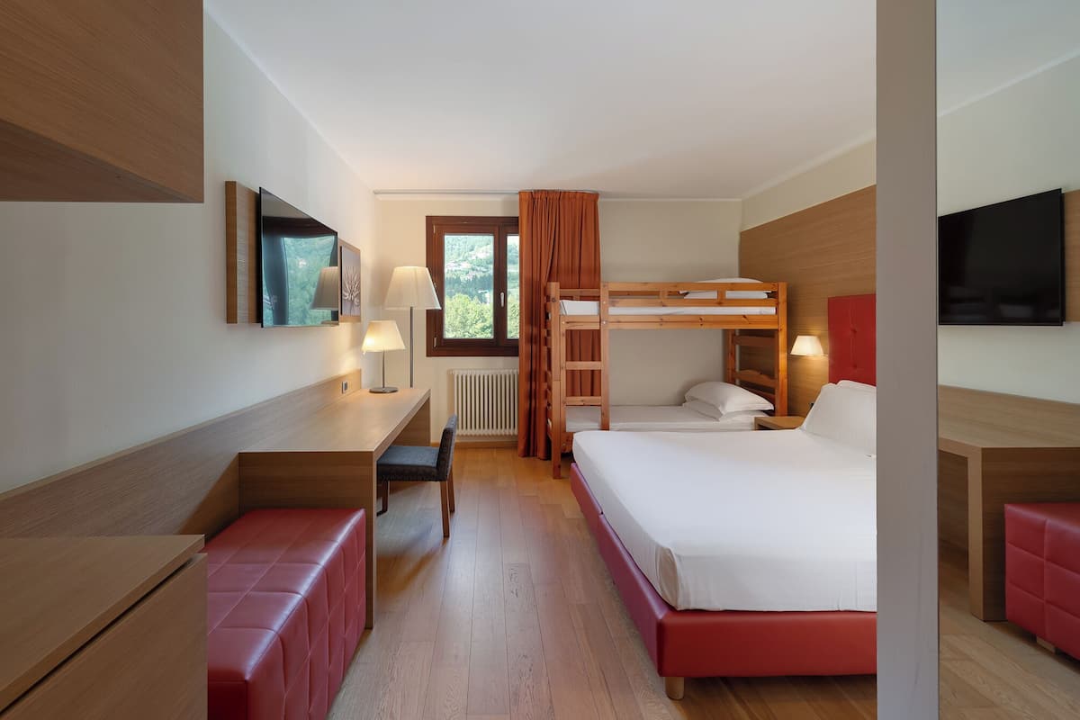 Une chambre d’hôtel avec un lit superposé, un bureau avec une chaise, un banc rouge, une télévision et une fenêtre avec des rideaux.
