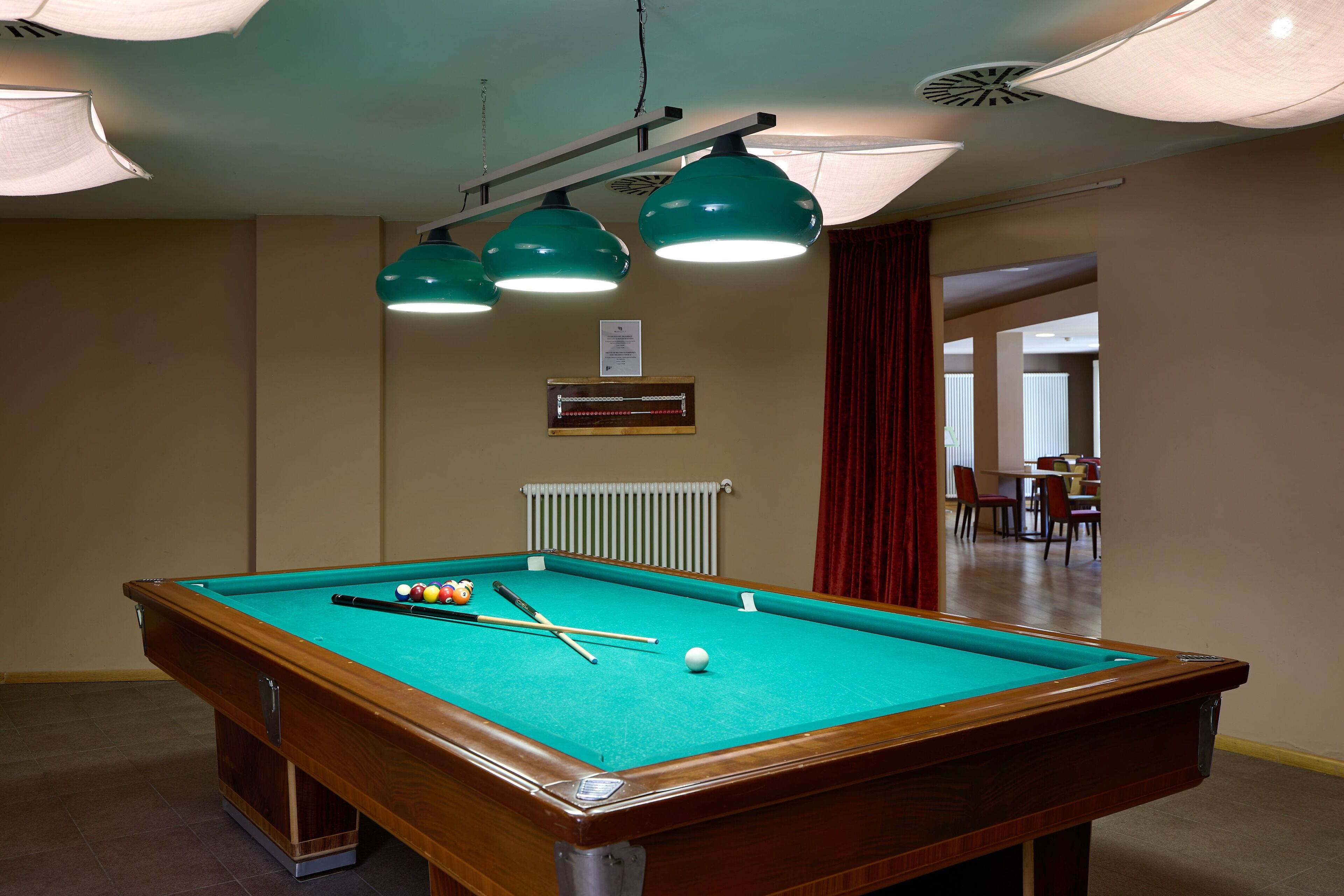 A pool table with green felt, surrounded by walls and a ceiling with green pendant lights.