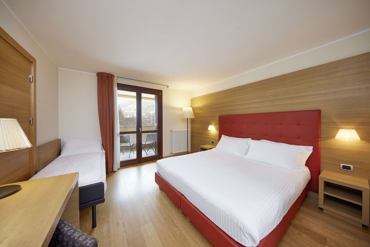 Une chambre d’hôtel avec un grand lit, un plus petit lit, un bureau et une chaise. Il y a une fenêtre avec des rideaux, un radiateur et une table de chevet avec une lampe.