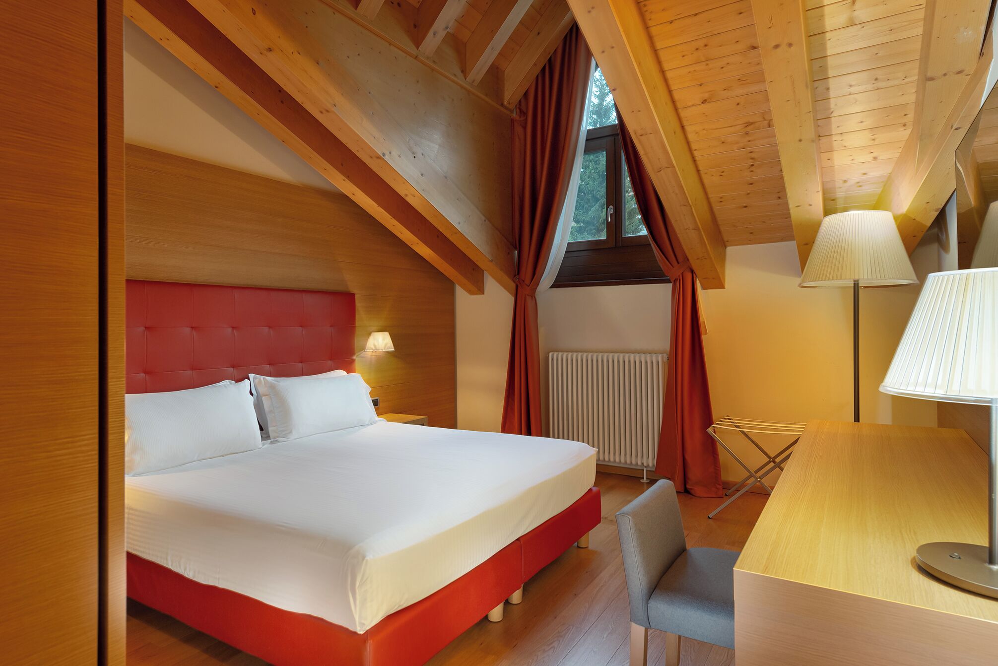 A bedroom with a red headboard, a bed with white linens, a bedside lamp, a radiator, and a desk with a chair.
