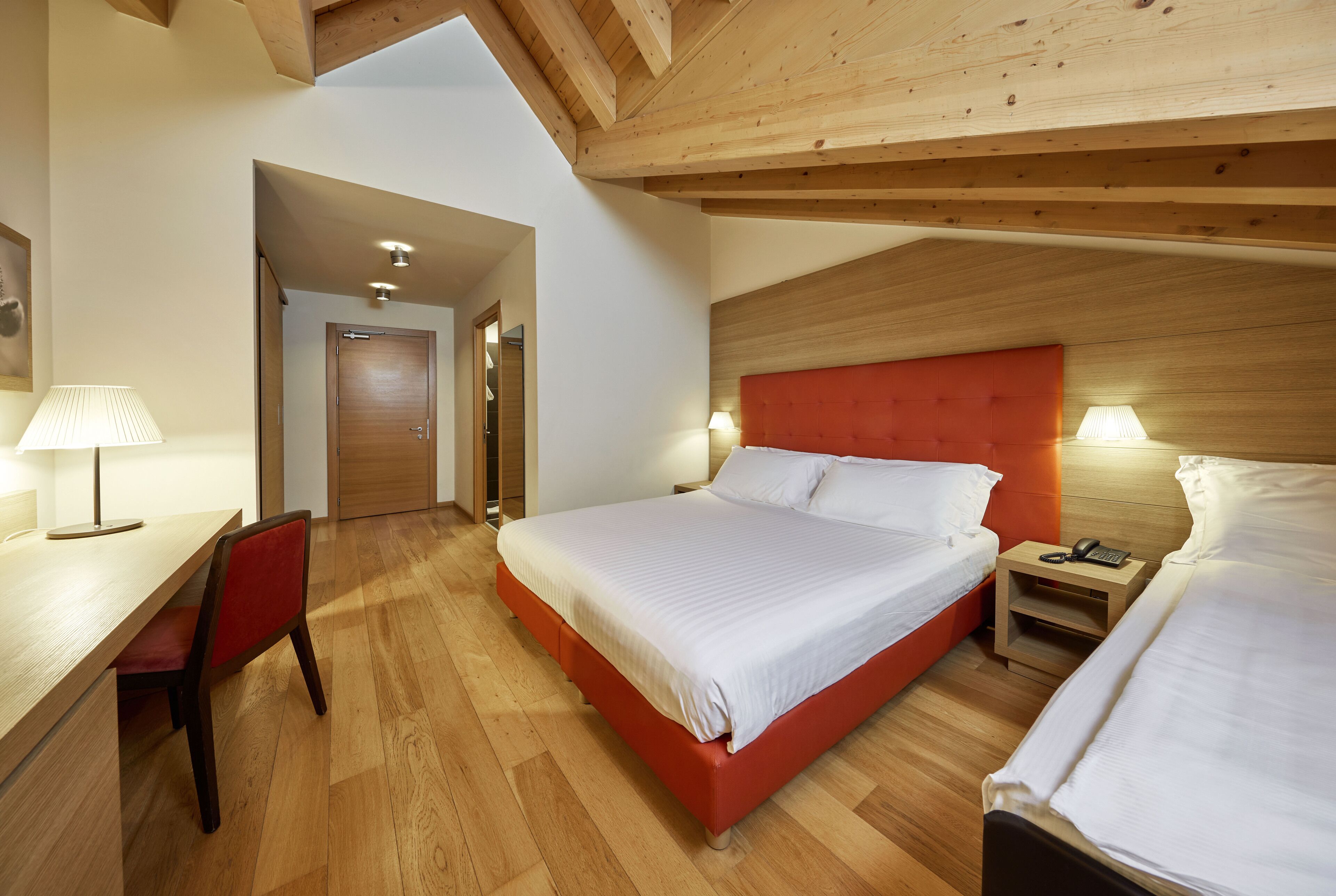 A hotel room with a wooden ceiling, a bed with white bedding, a red upholstered headboard, a bedside table with a lamp, and a desk with a chair.
