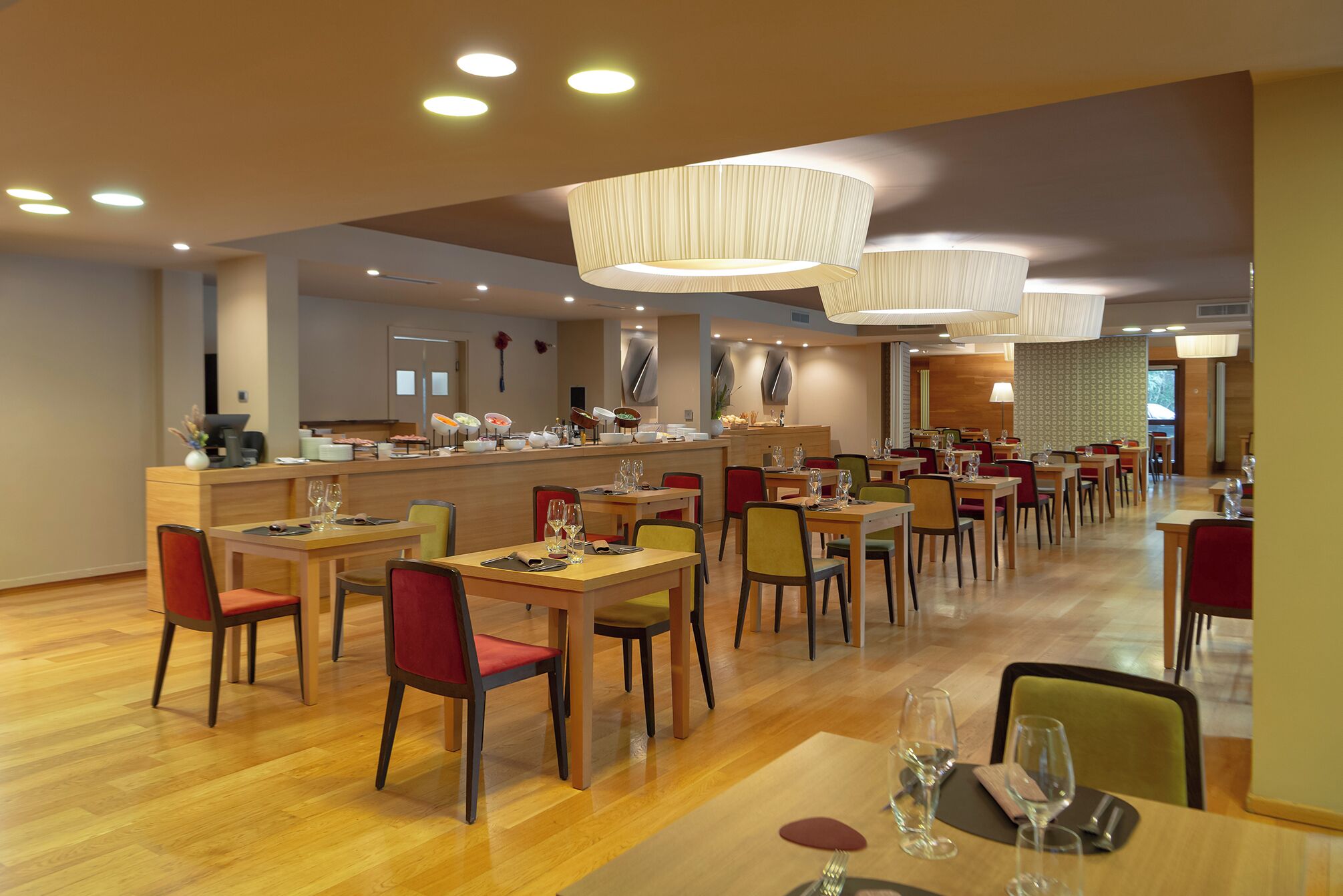 Un restaurante moderno con mesas y sillas de madera, una zona de buffet e iluminación colgante.