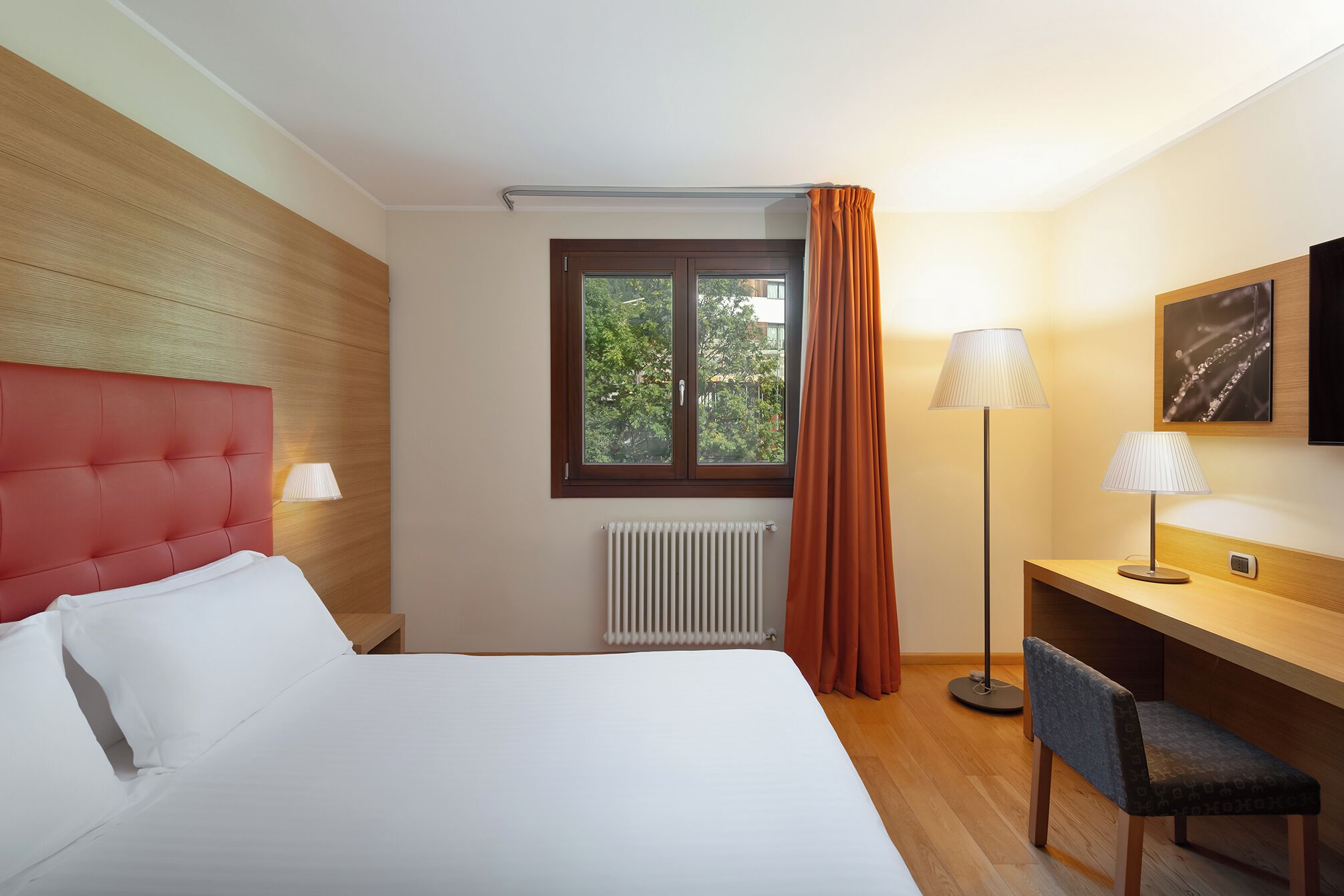 A hotel room with a bed, a desk, a chair, a radiator, a window with curtains, and a lamp.