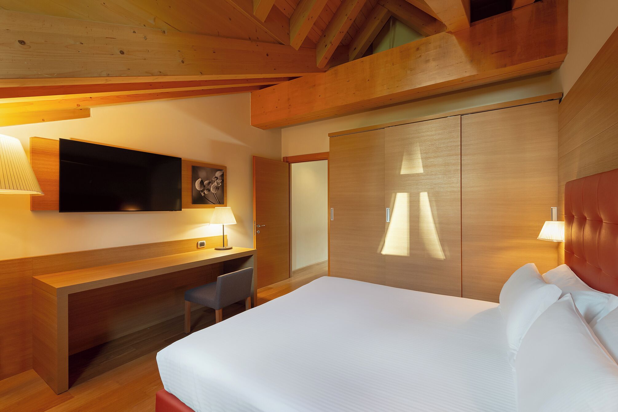 A modern hotel room with a large bed, a desk, a chair, a TV, and a wardrobe.