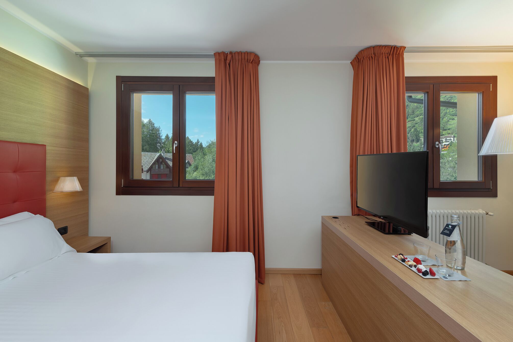 Habitación de hotel con cama, televisor, escritorio con una botella y vasos, y dos ventanas con cortinas.