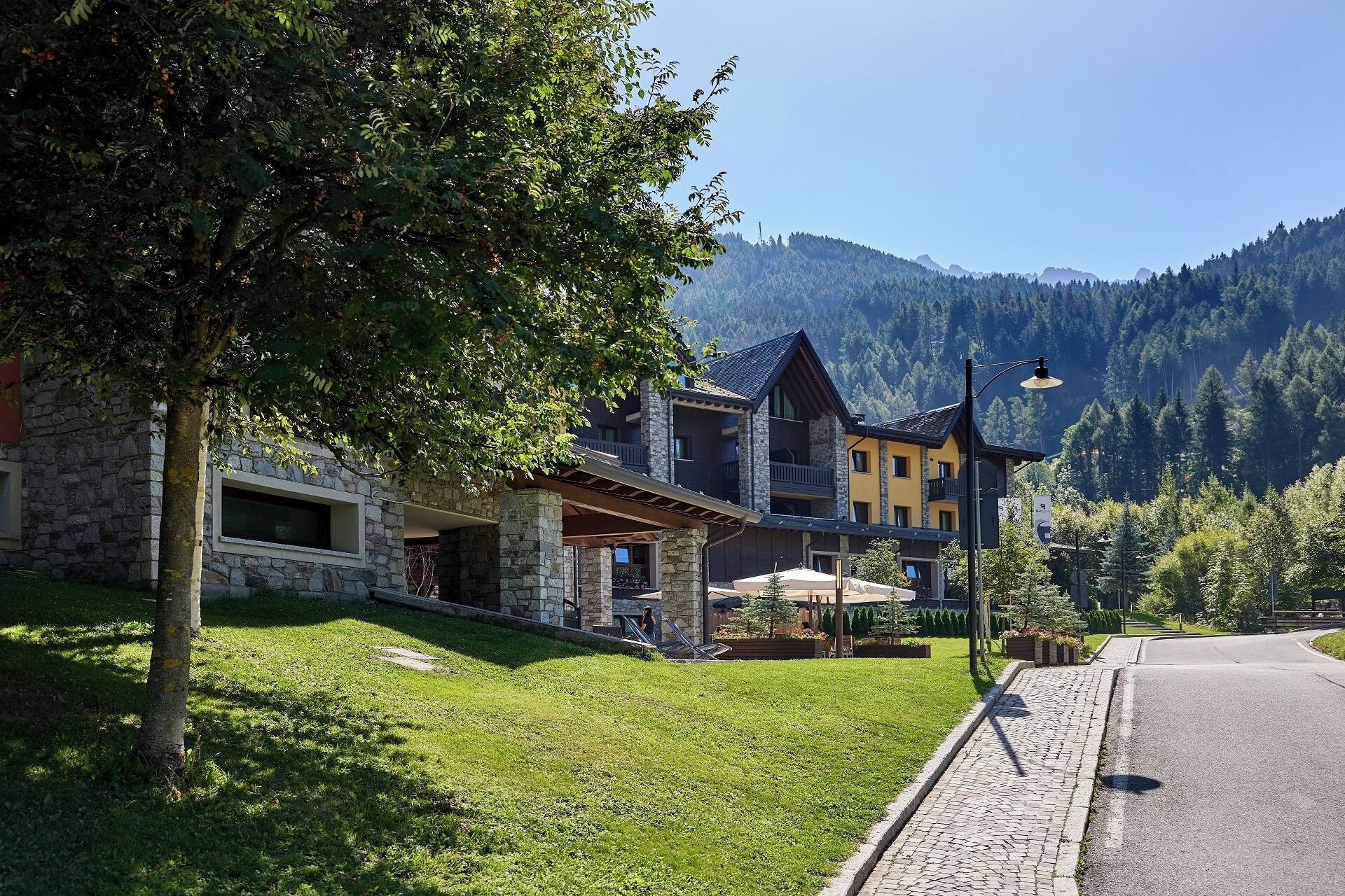 Un immeuble à plusieurs étages, avec une façade en pierre et en bois, une allée pavée et une vue sur les montagnes en arrière-plan.