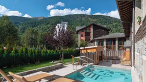 Área de piscina com espreguiçadeiras, deck de madeira e montanhas ao fundo.