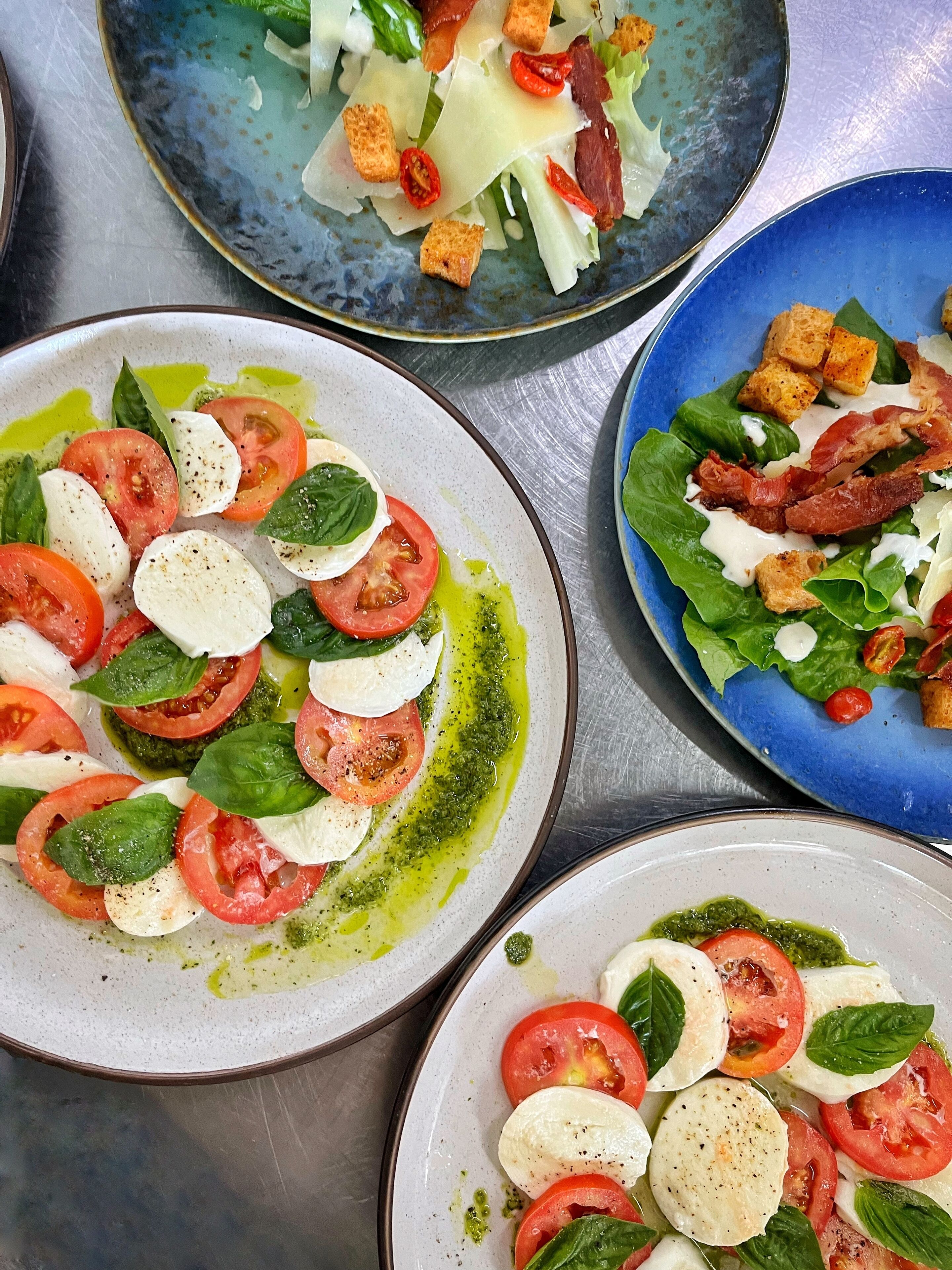 Trois assiettes de salade caprese, composées de tomates, de mozzarella et de basilic, avec des croûtons, le tout disposé sur une surface métallique.