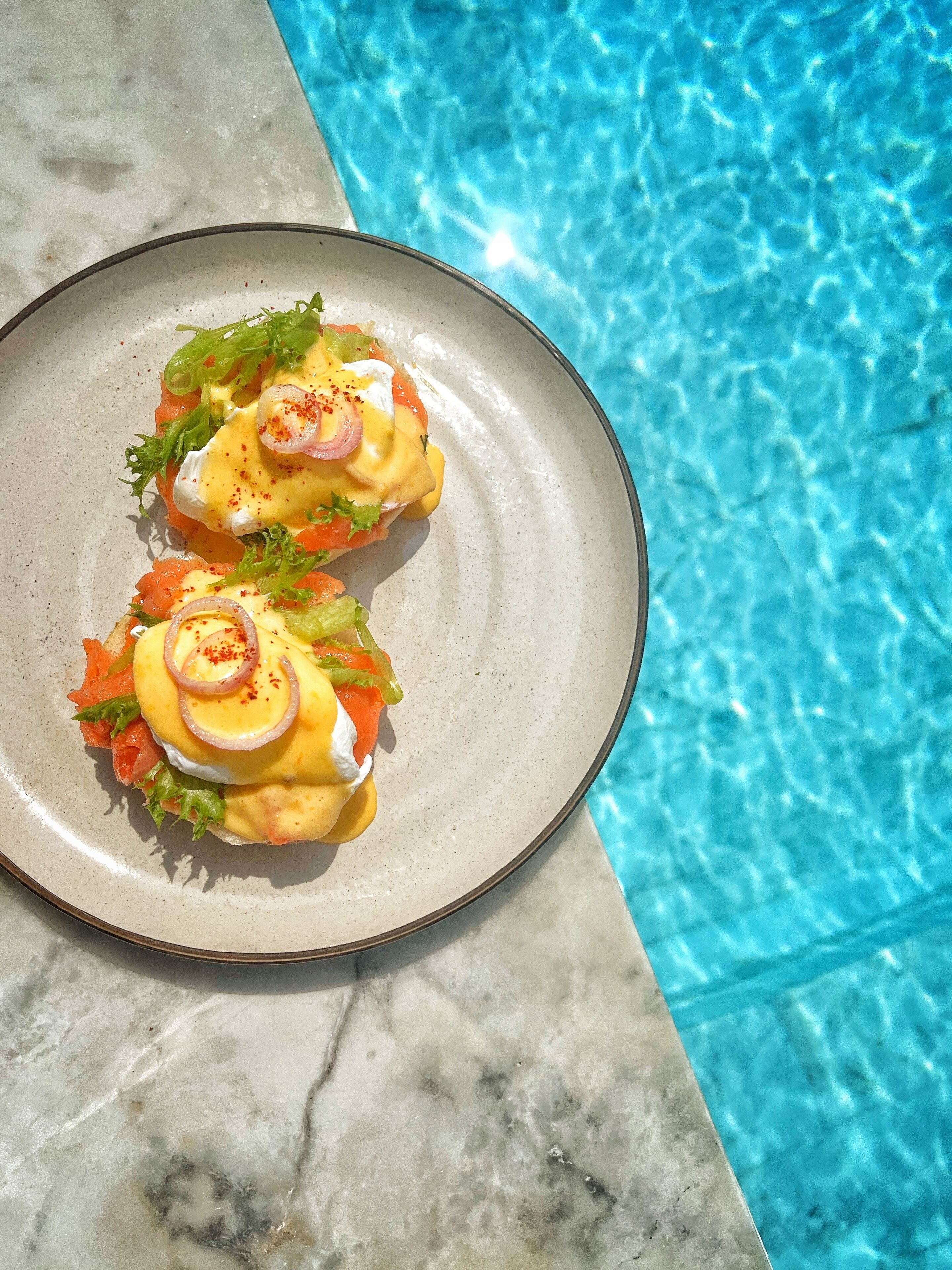 Un plato de huevos benedictinos con salsa holandesa, adornado con hojuelas de chile, sobre una superficie de mármol junto a una piscina.