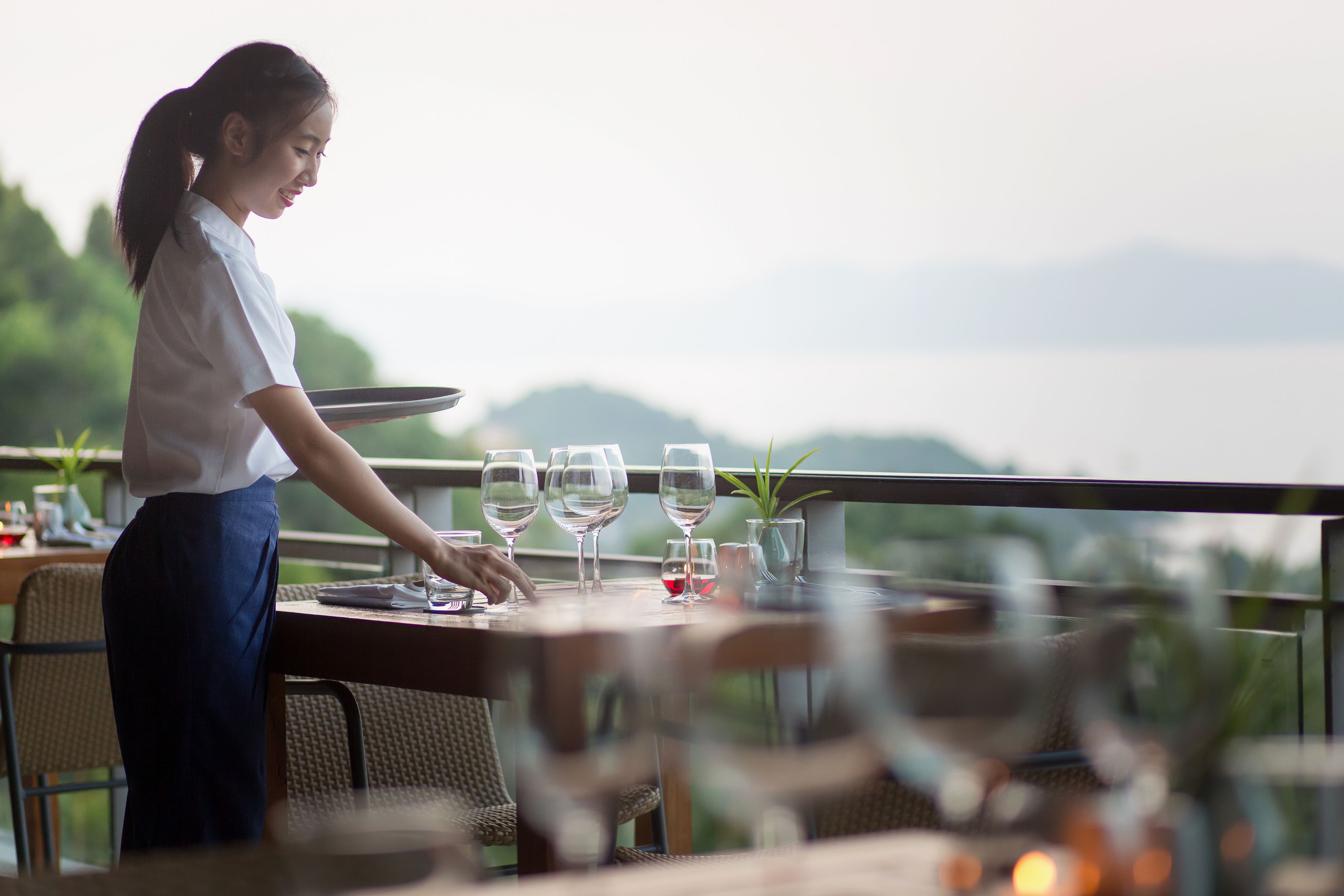 Une personne prépare une table avec des verres à vin et une plante sur un balcon donnant sur un paysage montagneux.