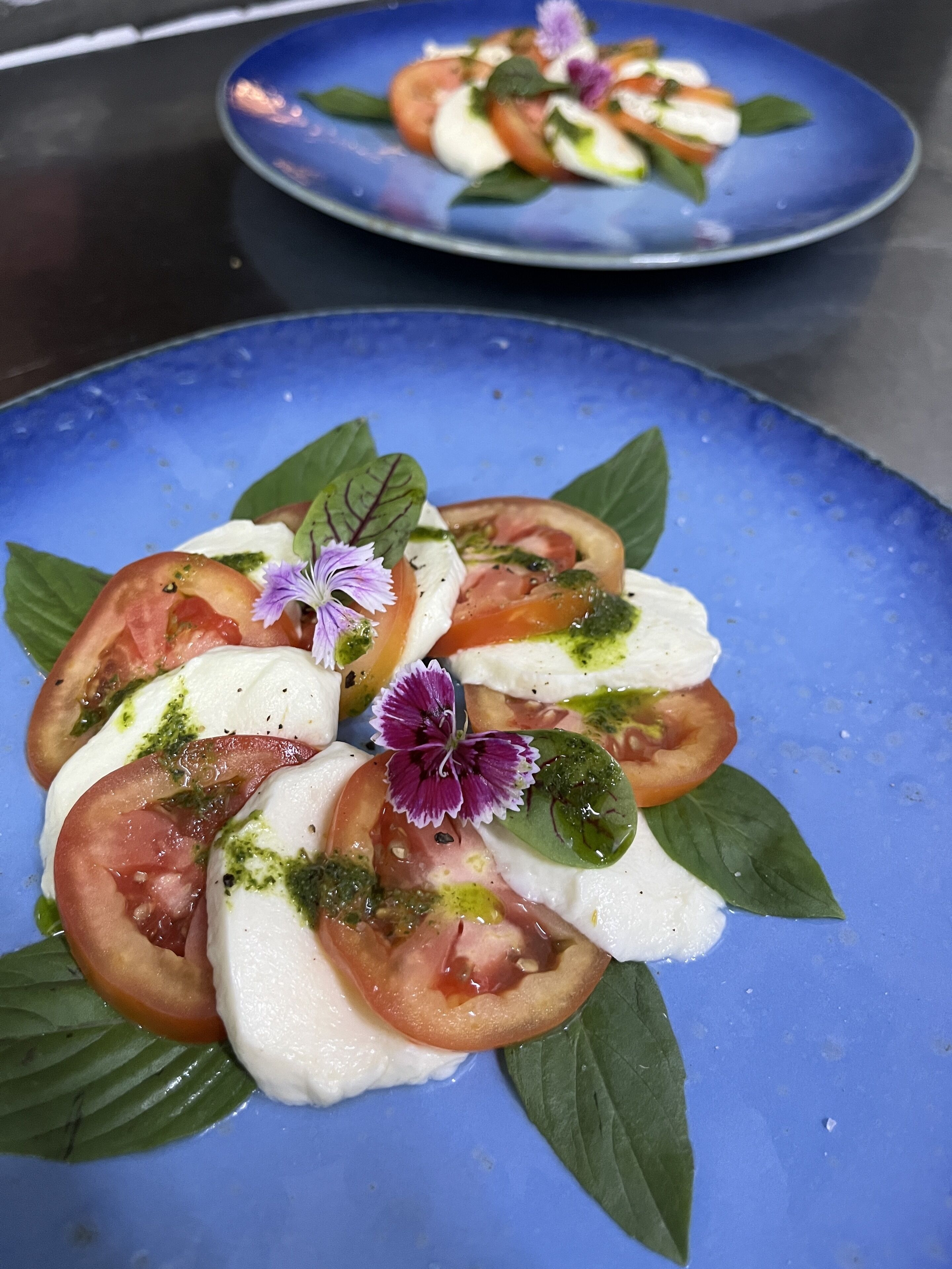 A blue plate with a tomato and mozzarella salad garnished with basil leaves and a purple flower.