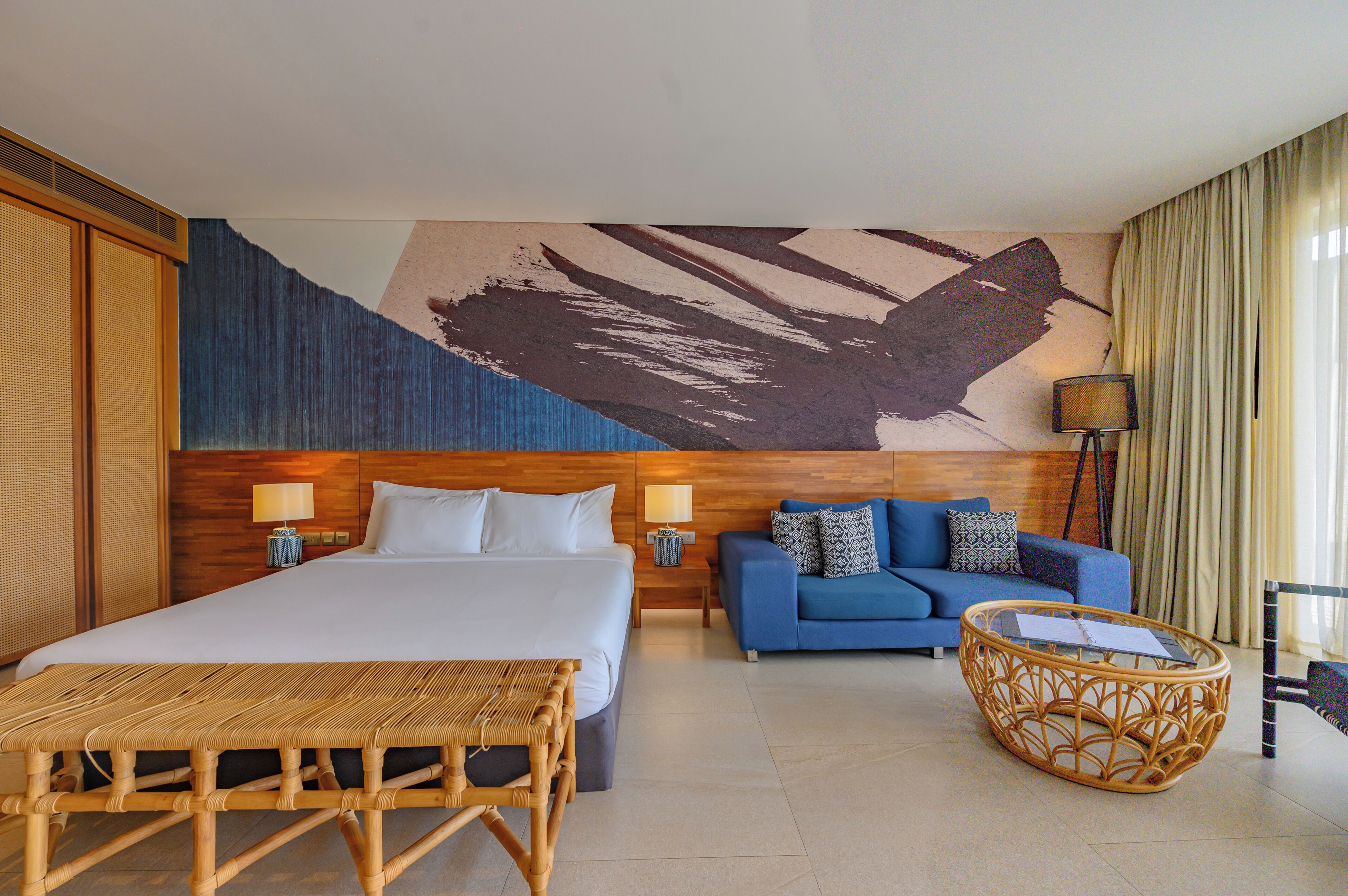 A hotel room with a large bed, a blue sofa, a wicker coffee table, and a wall painting.