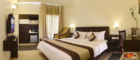 A hotel room with a large bed, a desk with a computer, a small refrigerator, and a TV.