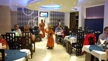 Un restaurante con mesas redondas, un escenario con artistas y una pantalla de televisión al fondo.