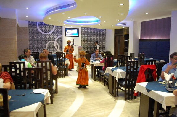 A restaurant with round tables, a stage with performers, and a TV screen in the background. - Hotel Taj Resorts (Agra)