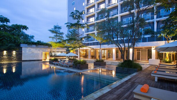 A pool area with lounge chairs and umbrellas, surrounded by a building with multiple balconies. - Delonix Hotel Karawang (Karawang)