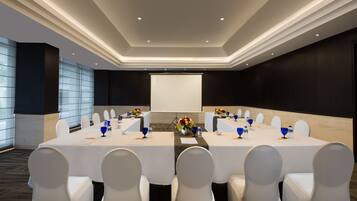 A conference room with a large table, white chairs, a projector screen, and floral centerpieces.