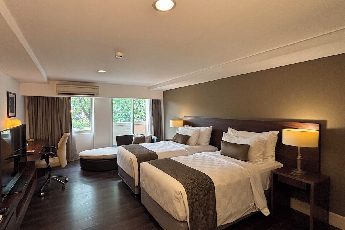 Quarto de hotel com duas camas, uma escrivaninha, uma cadeira, uma televisão e uma janela com cortinas.