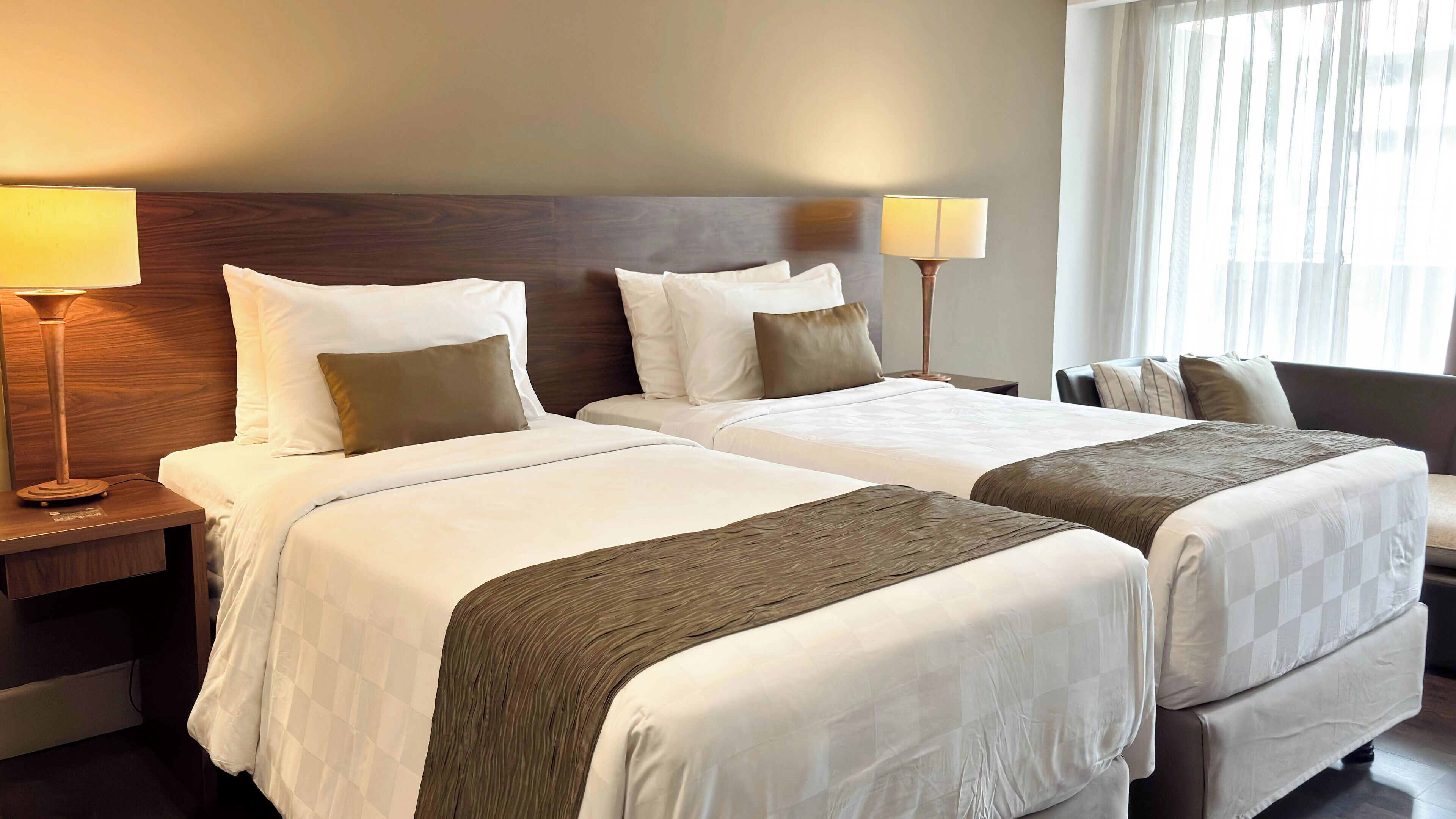 A hotel room with two beds, each with white linens and brown throw blankets. There are two bedside tables with lamps and a large window with sheer curtains.