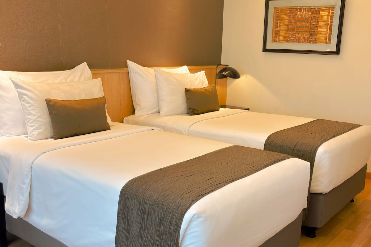 A hotel room with two beds, each with white linens and brown throw blankets. There are two bedside tables with lamps, and a framed artwork on the wall.