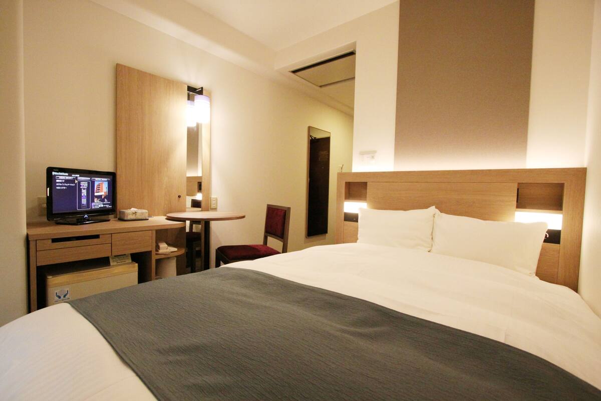 A hotel room with a bed, a desk with a computer, a chair, and a small table.