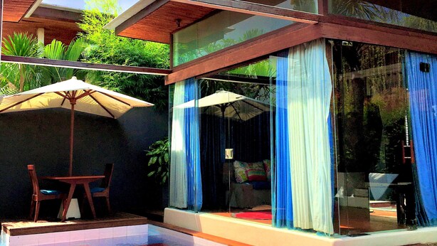A modern outdoor pool area with a wooden deck, two umbrellas, and a glass-enclosed space with blue curtains. - Kiss Villas Bali (Seminyak)